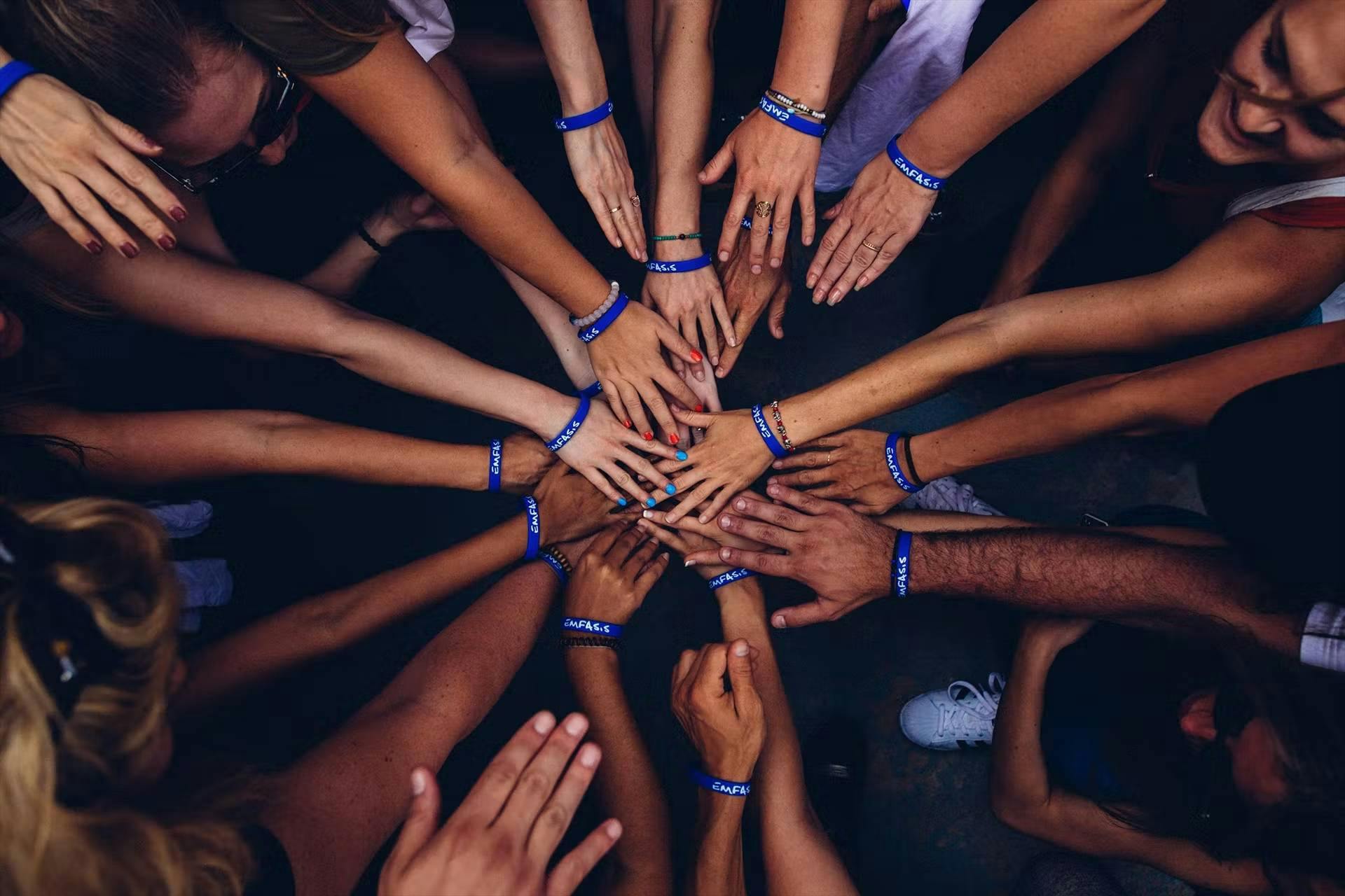 Dizaines de personnes en cercle qui tendant le bras pour rassembler leurs mains au milieu - ils portent un bracelet bleu 