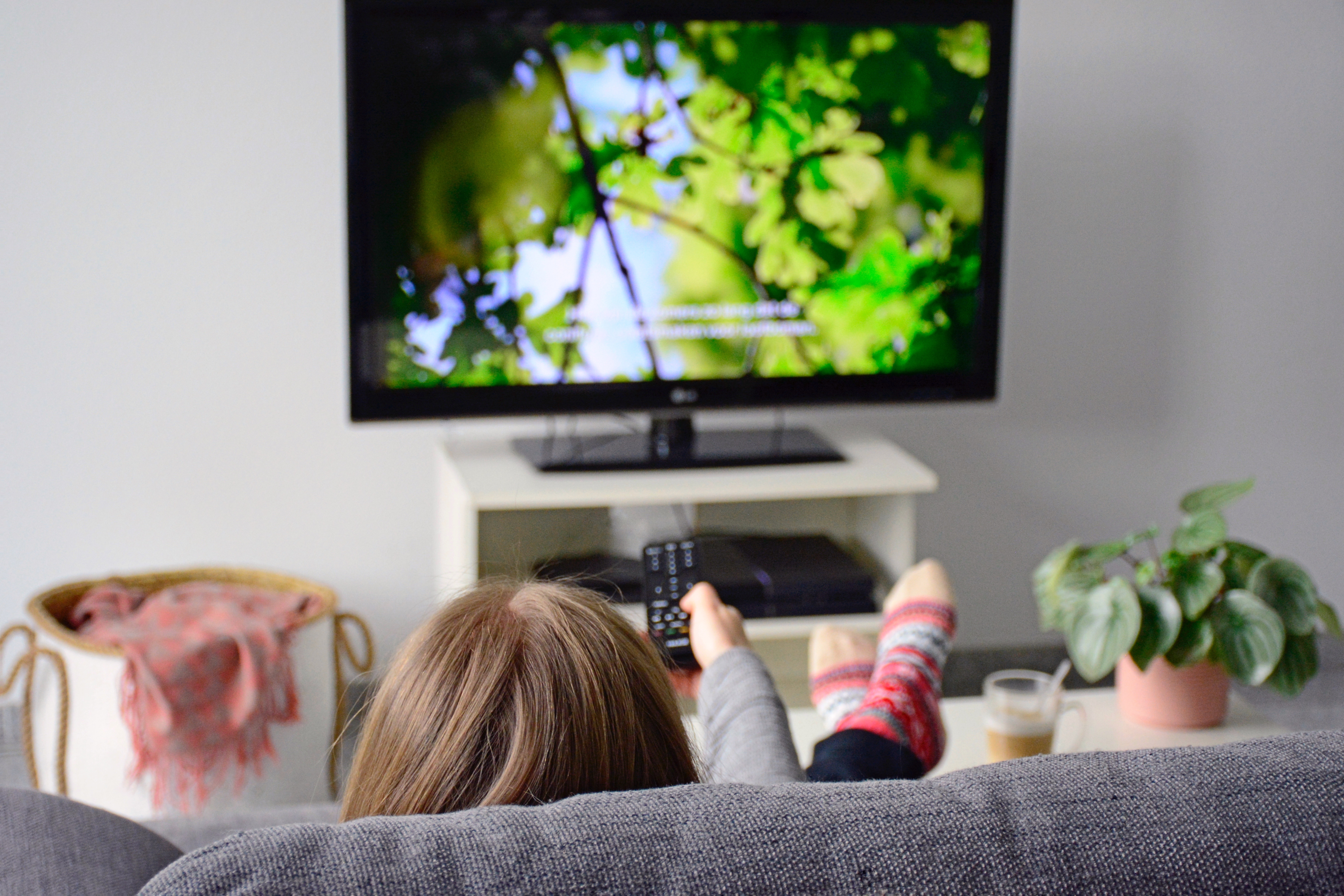 A woman sitting on the sofa with her feet up in front of the TV. The woman points the remote towards the TV as the picture of a forest appears.