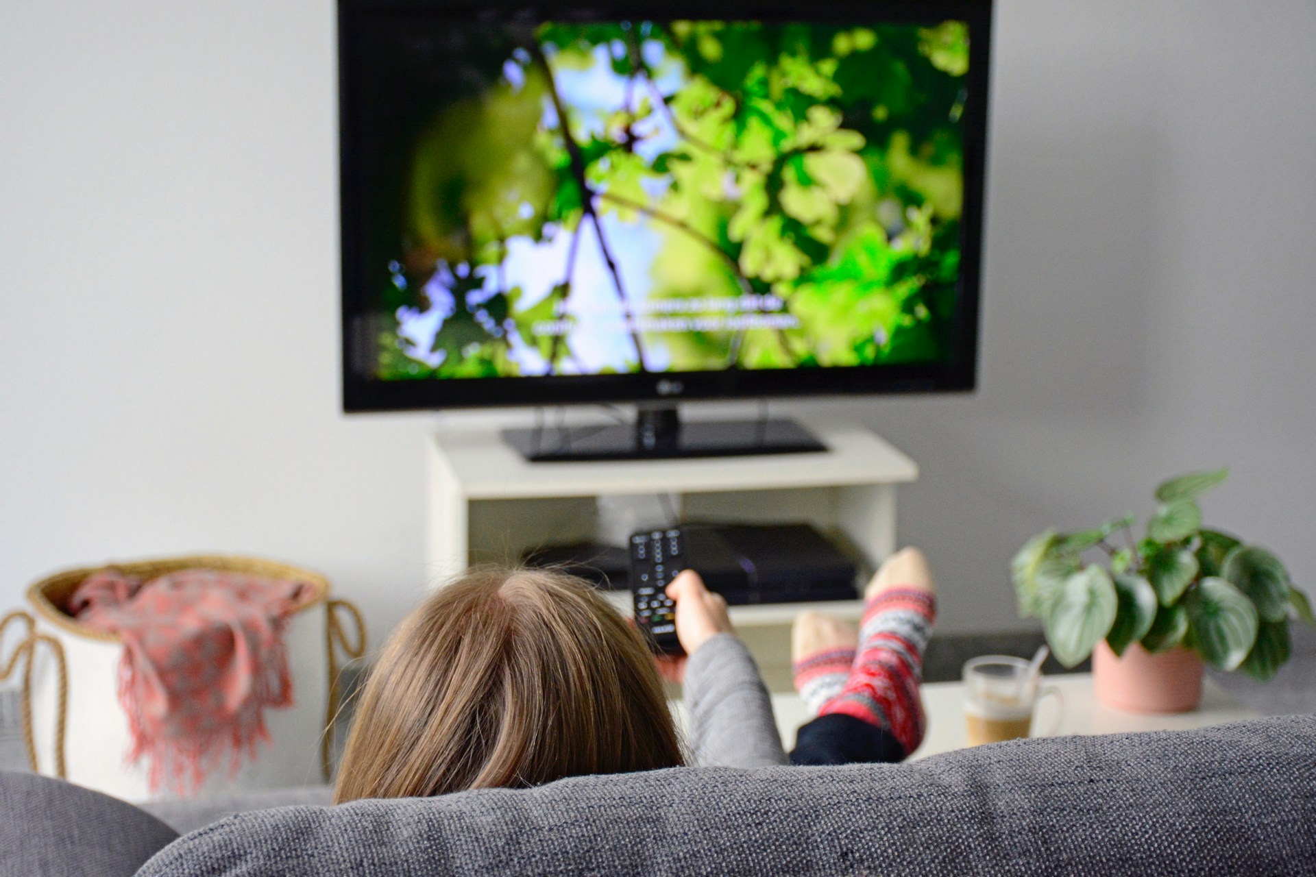 A woman sitting on the sofa with her feet up in front of the TV. The woman points the remote towards the TV as the picture of a forest appears.