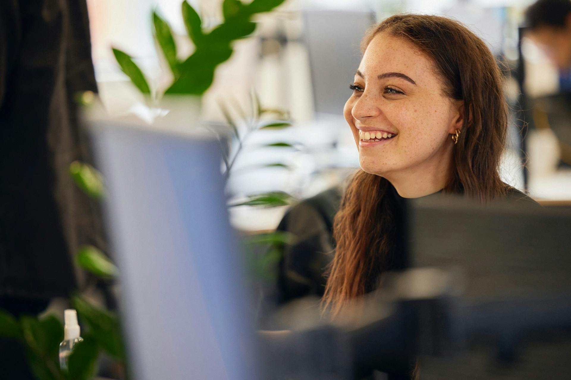 A redheaded person smiles in a busy office in this header image for Meltwater's customer story about CCGroup.