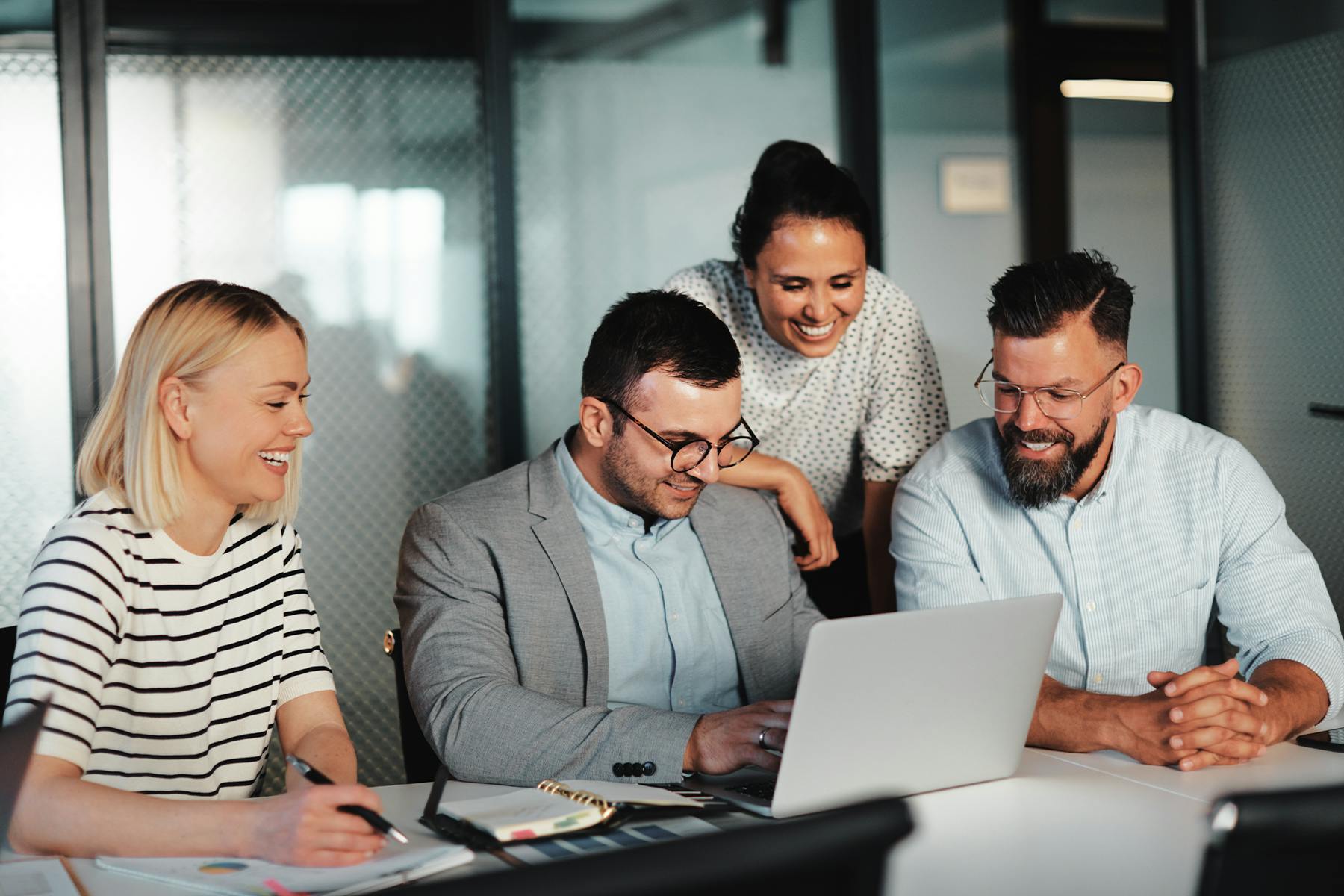 Four people in a meeting room look at a laptop screen while smiling