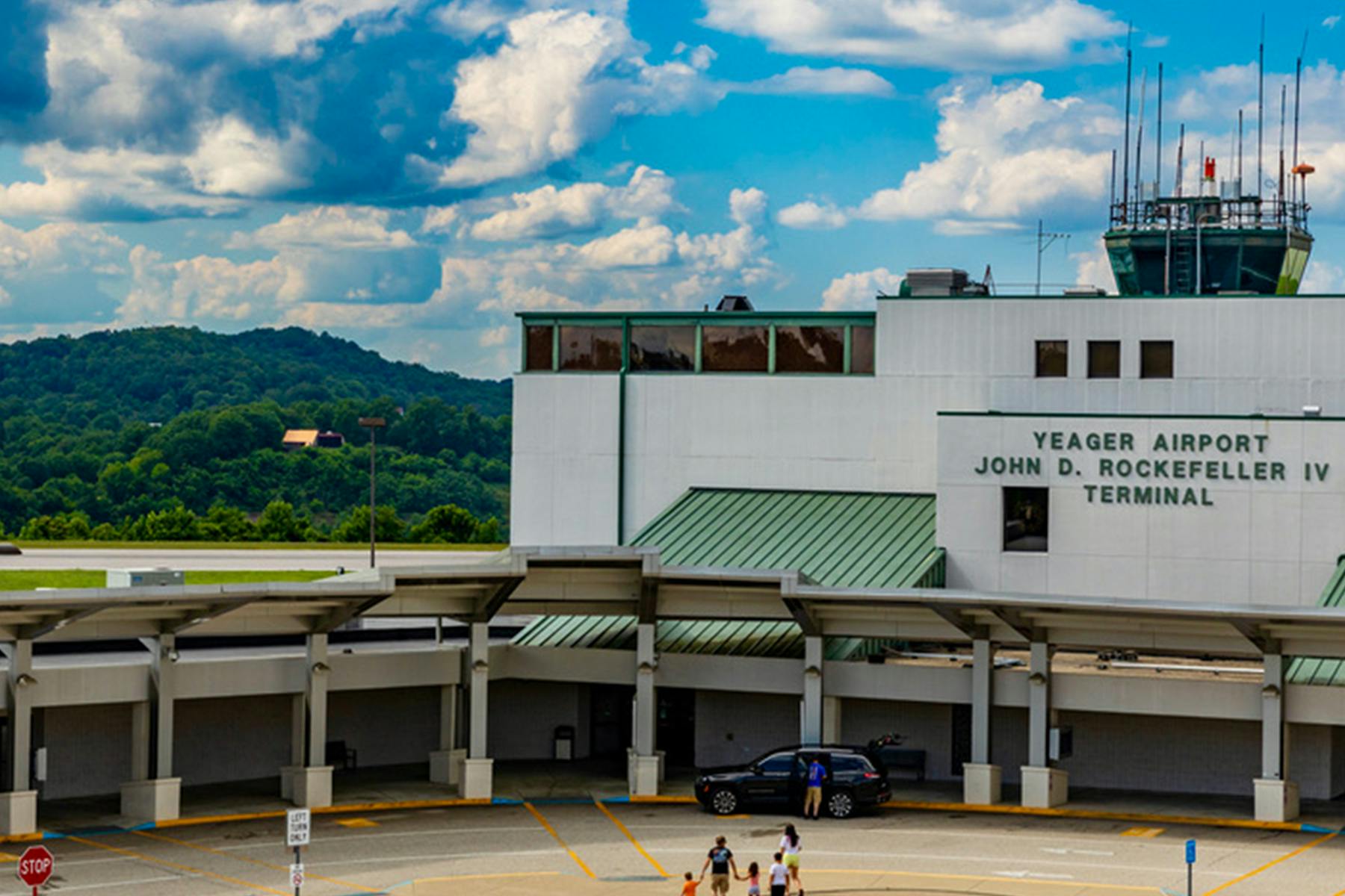 Exterior view of Yeager Airport’s John D. Rockefeller IV Terminal, a white building with green metal roofs and a control tower, set against tree-covered hills and a partly cloudy sky.