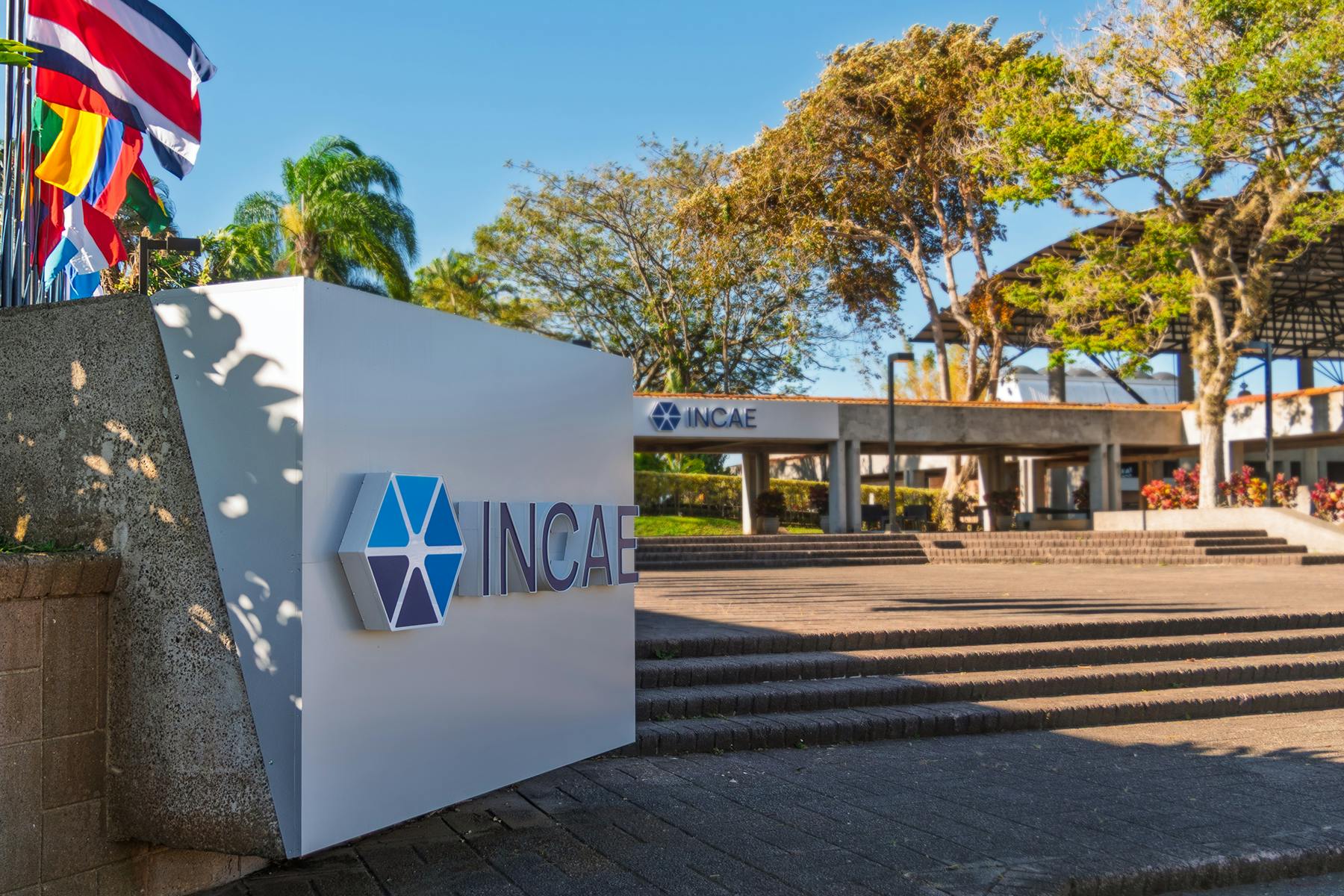 Exterior view of INCAE Business School campus with a large INCAE sign in the foreground, palm trees and flags to the left, and the main building entrance in the background under a clear blue sky.