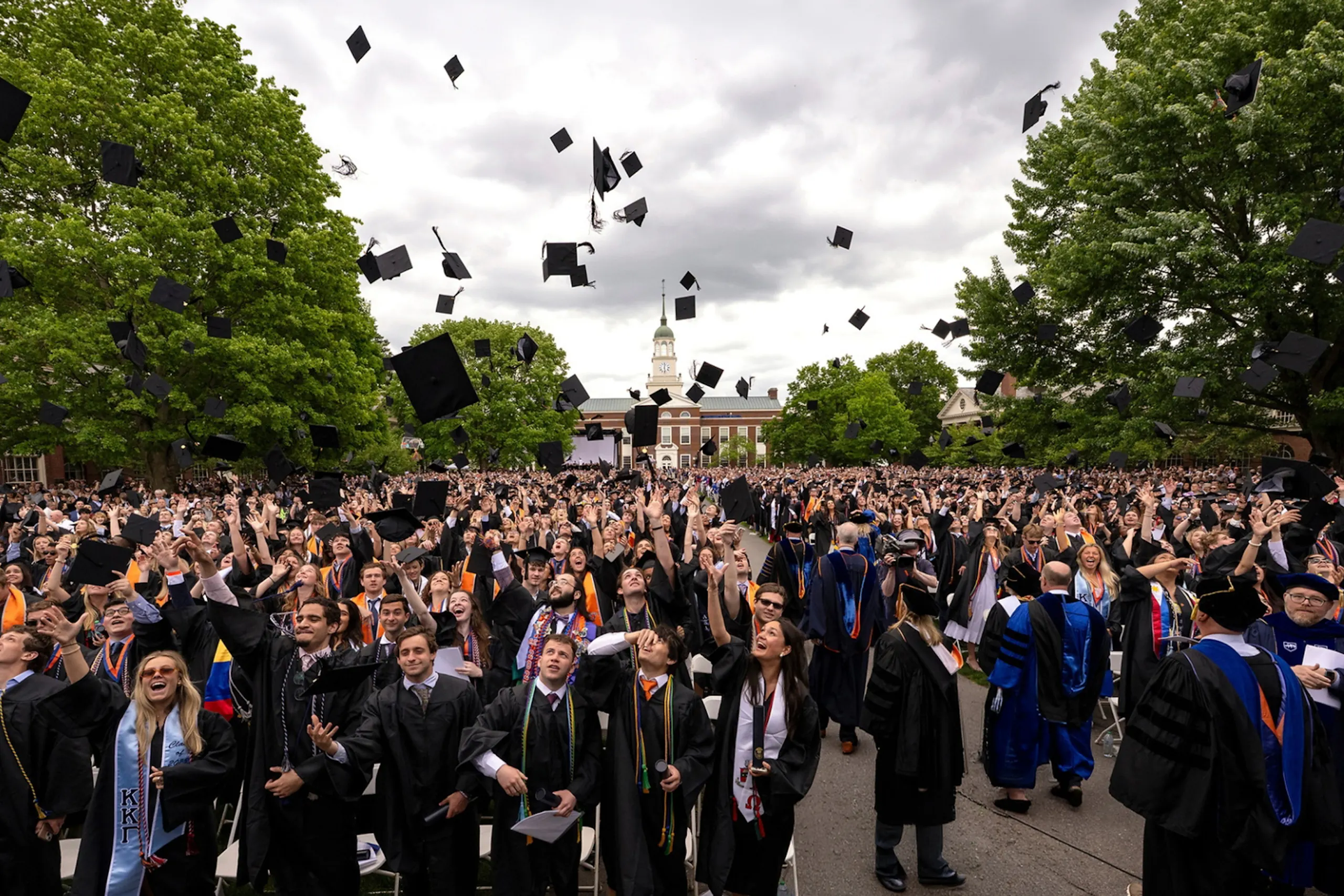 Hundreds of Bucknell University graduates in black caps and gowns celebrate at an outdoor commencement ceremony, tossing their mortarboards into the air as a campus building and large trees frame the scene under a cloudy sky.