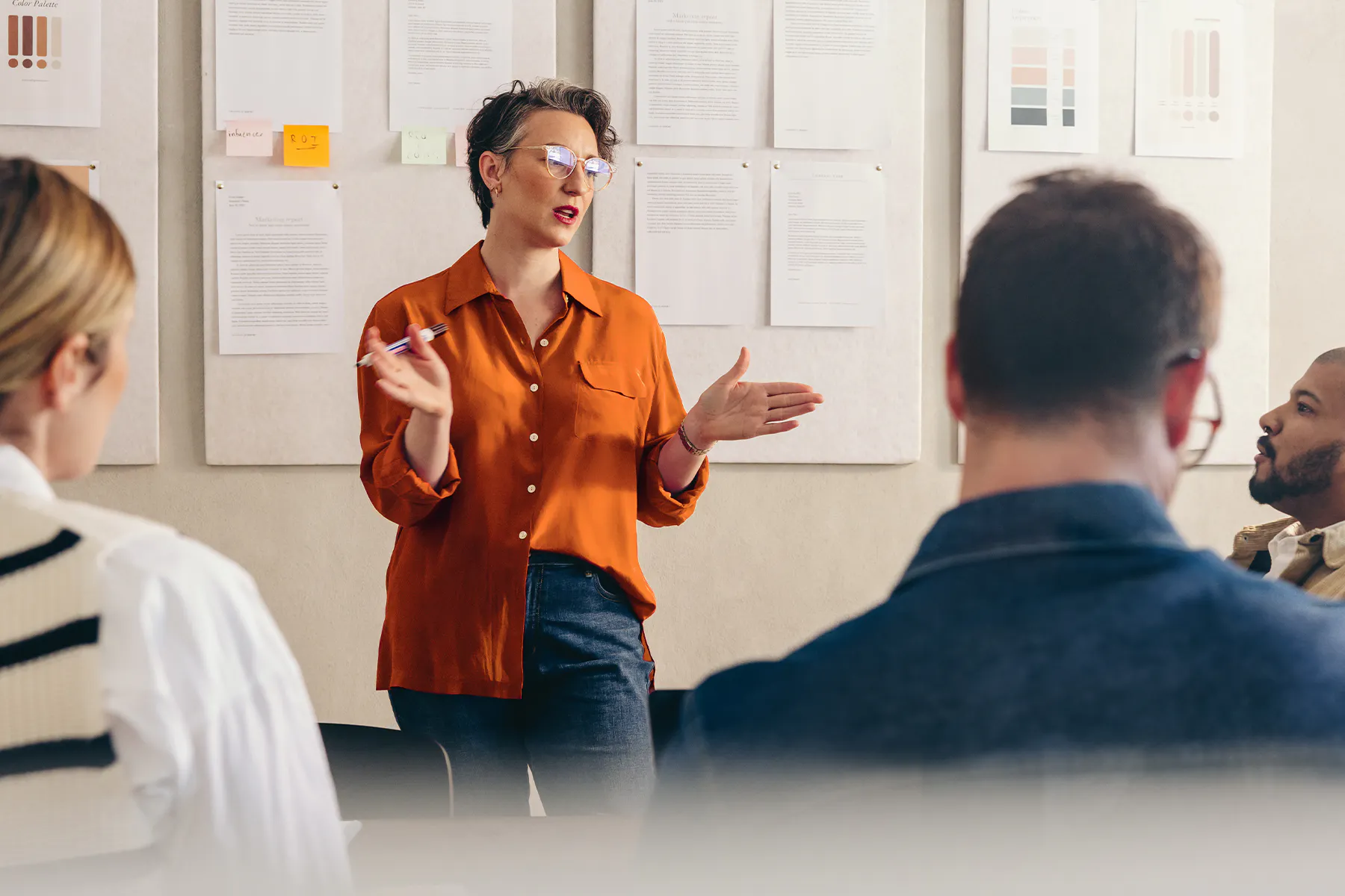 Professional presenting to colleagues during a team meeting, standing in front of a wall displaying reports, design mockups, and notes while discussing ideas and strategy in a collaborative office setting.