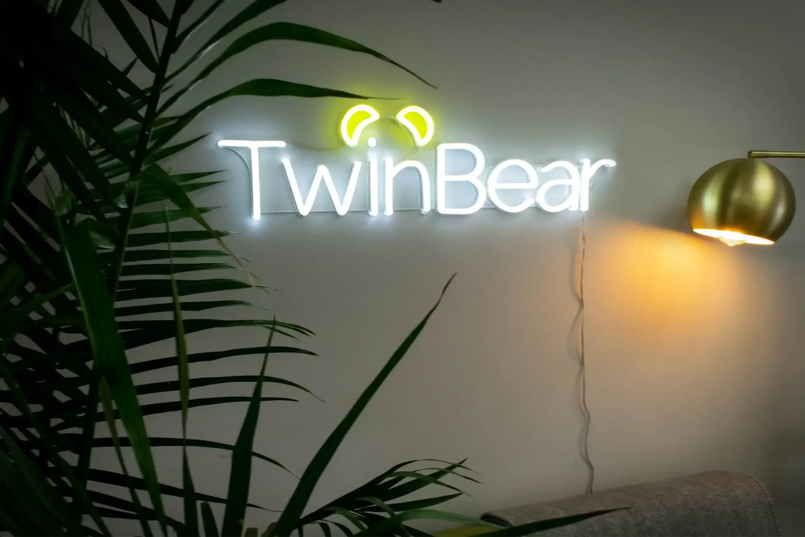 Illuminated TwinBear neon wall sign with stylized bear ears above the logo, mounted on a light-colored wall beside a gold floor lamp and surrounded by green indoor plants.