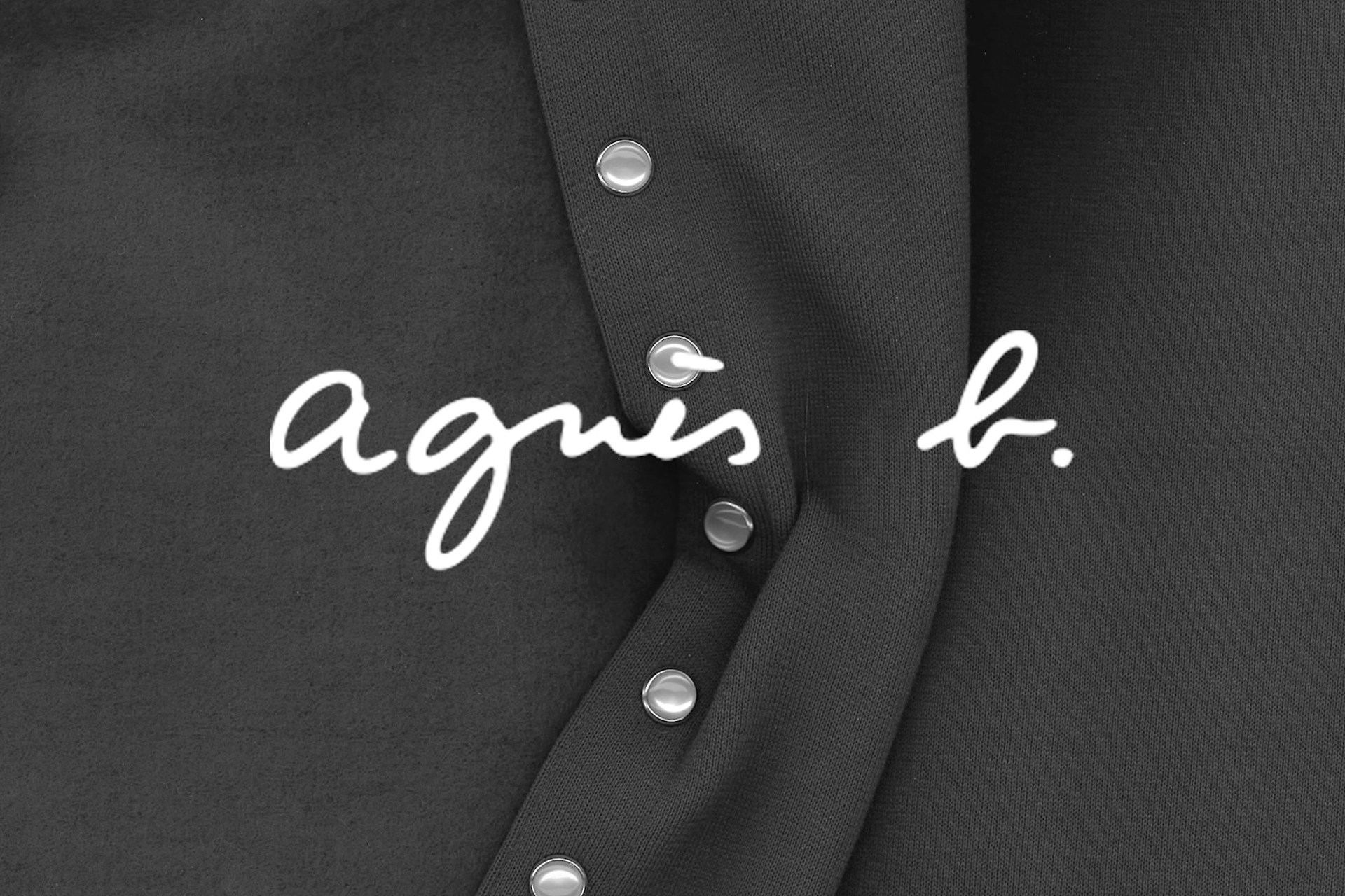 Logo Agnès b. écrit en blanc sur tissu noir avec des boutons