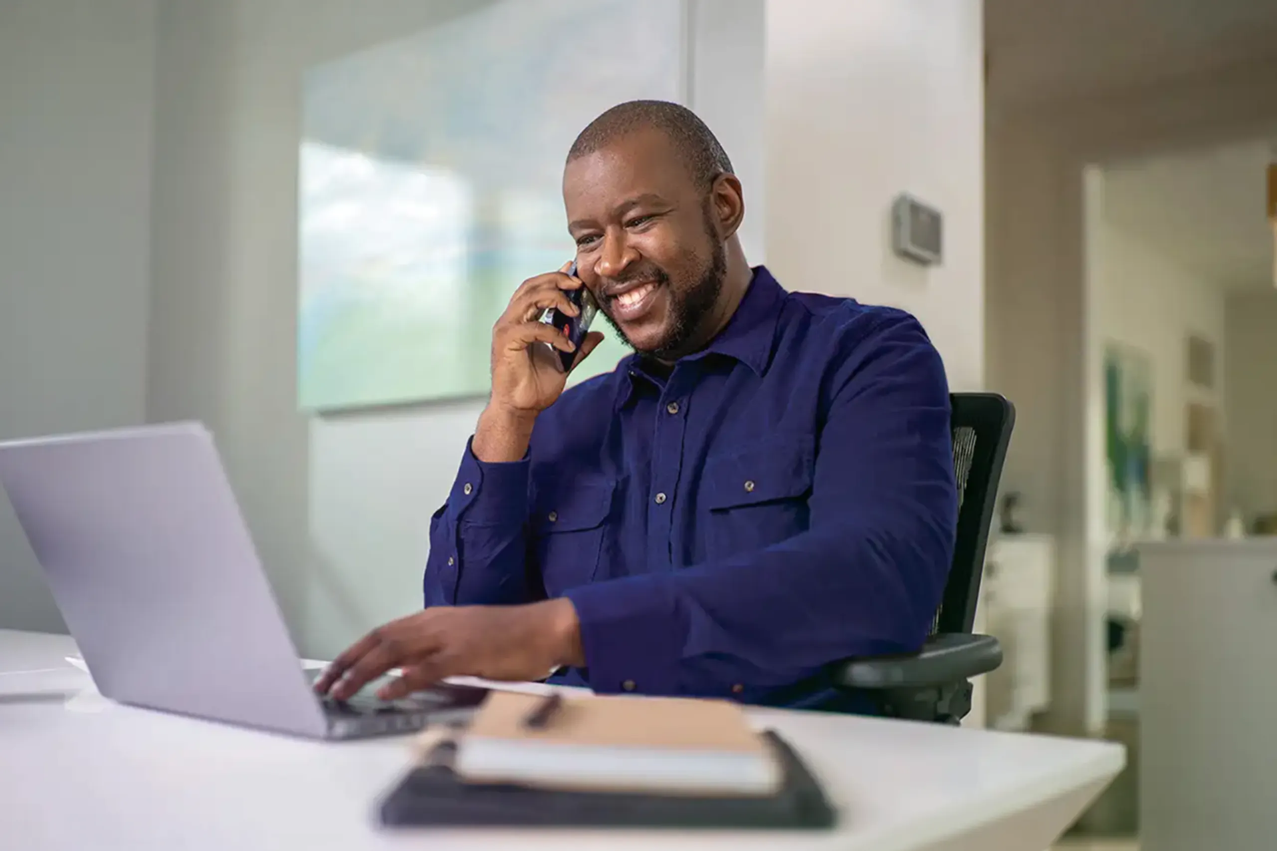 A man sitting in front of a laptop and speaking on a cellphone.