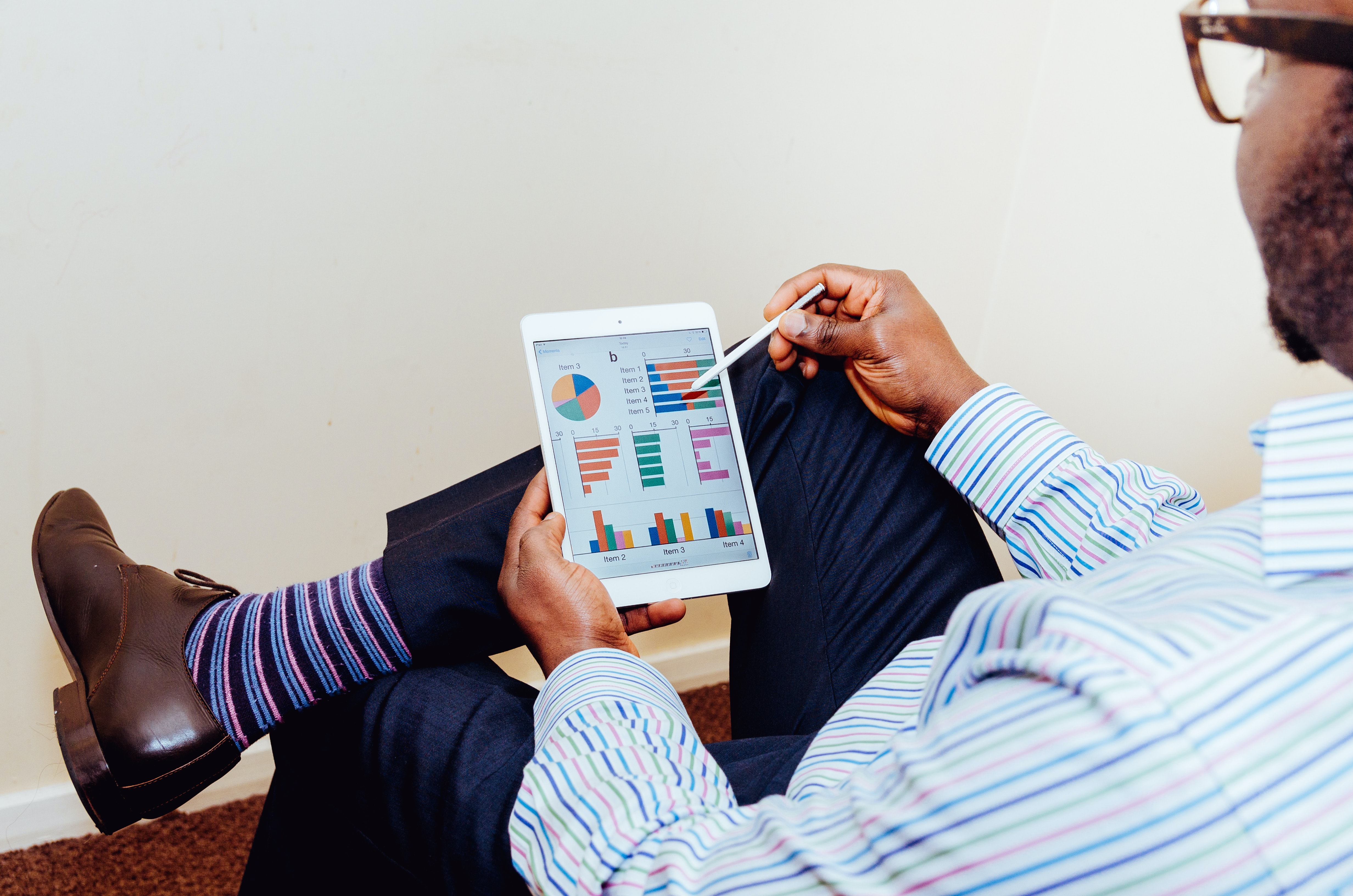 A man sitting in a chair and looking at a tablet screen. He is wearing striped purple socks, a striped shirt, and eyeglasses. The screen of the tablet shows various types of graphs.
