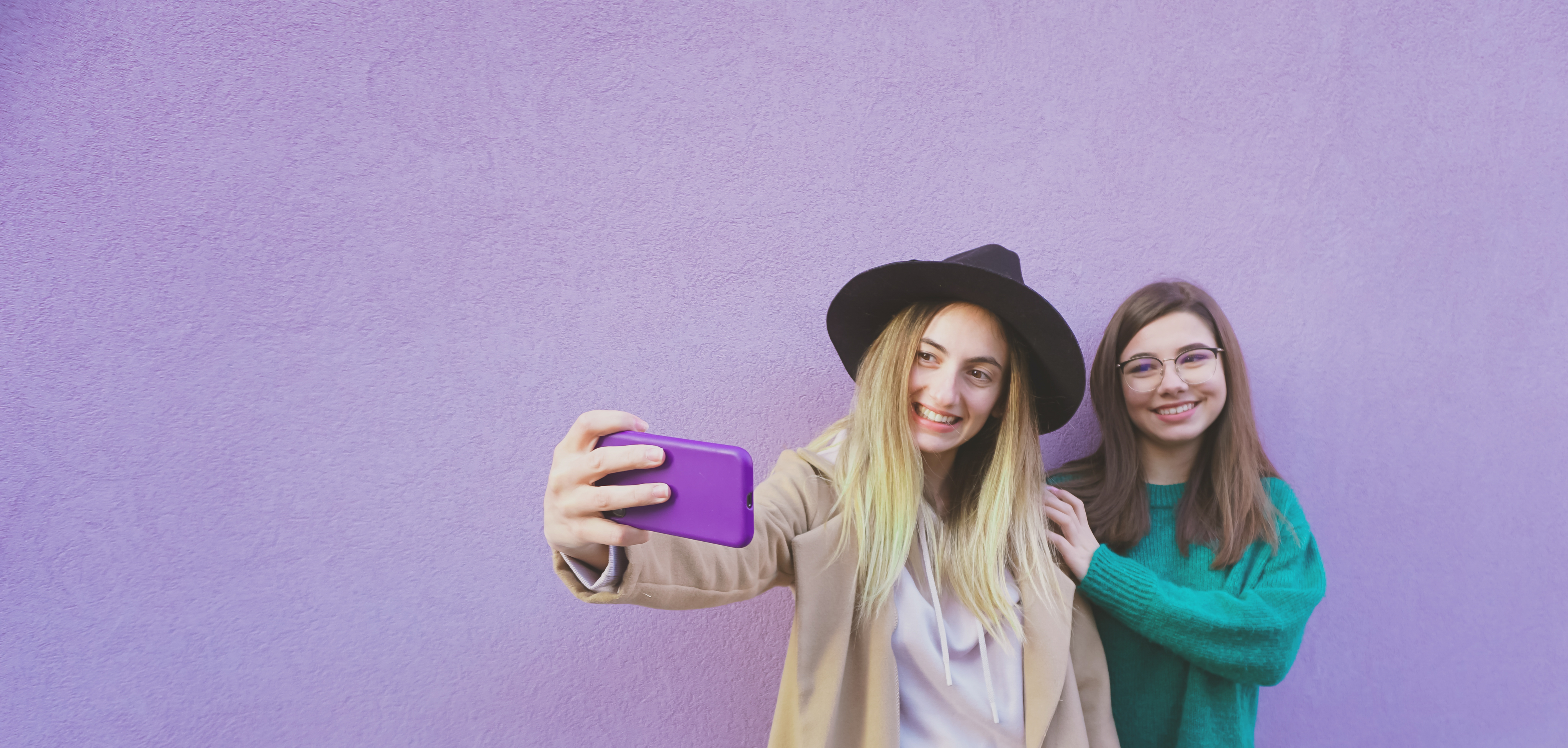 Two young girls, taking a selfie in front of a purple wall. 