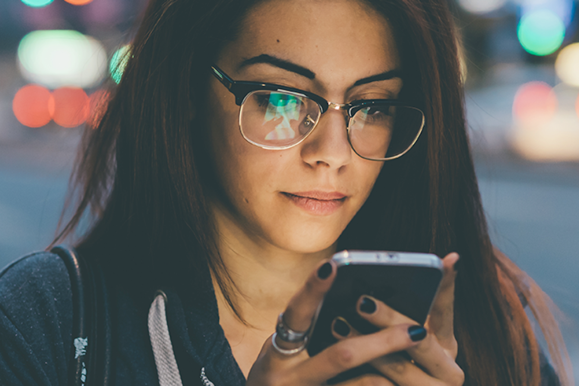 A young woman with brown hair and clear glasses looking at her phone screen. 