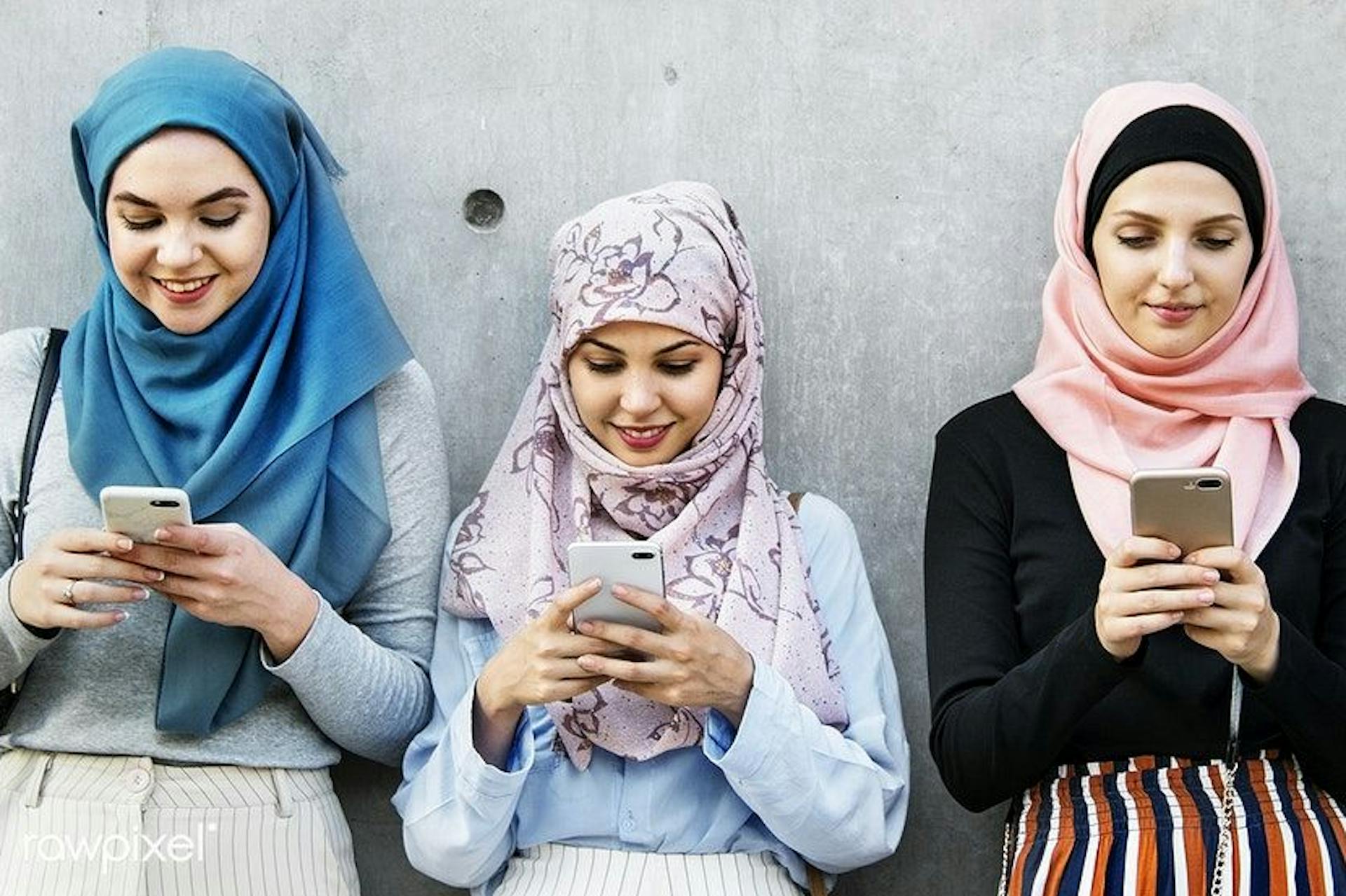 四个阿拉伯妇女靠在一堵灰色的墙上，看着她们的智能手机