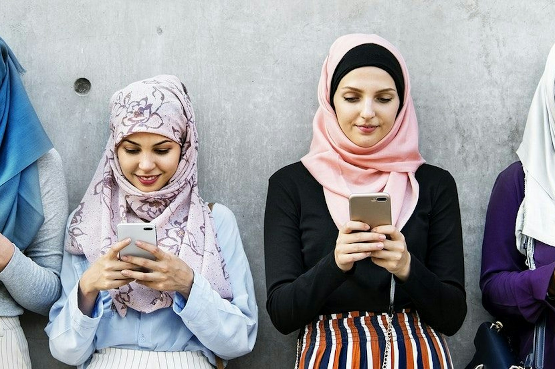 four arab women against a grey wall looking at their smartphones