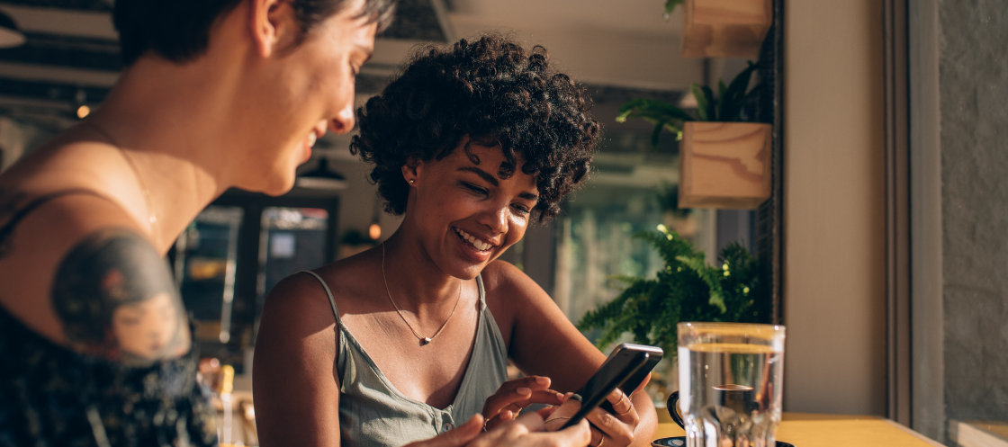 Two people sitting at a table looking at a smart-phone and smiling