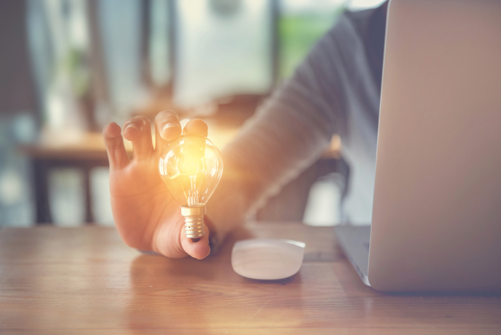 A woman at a desk with a laptop and mouse. In her hand is a lit lightbulb.