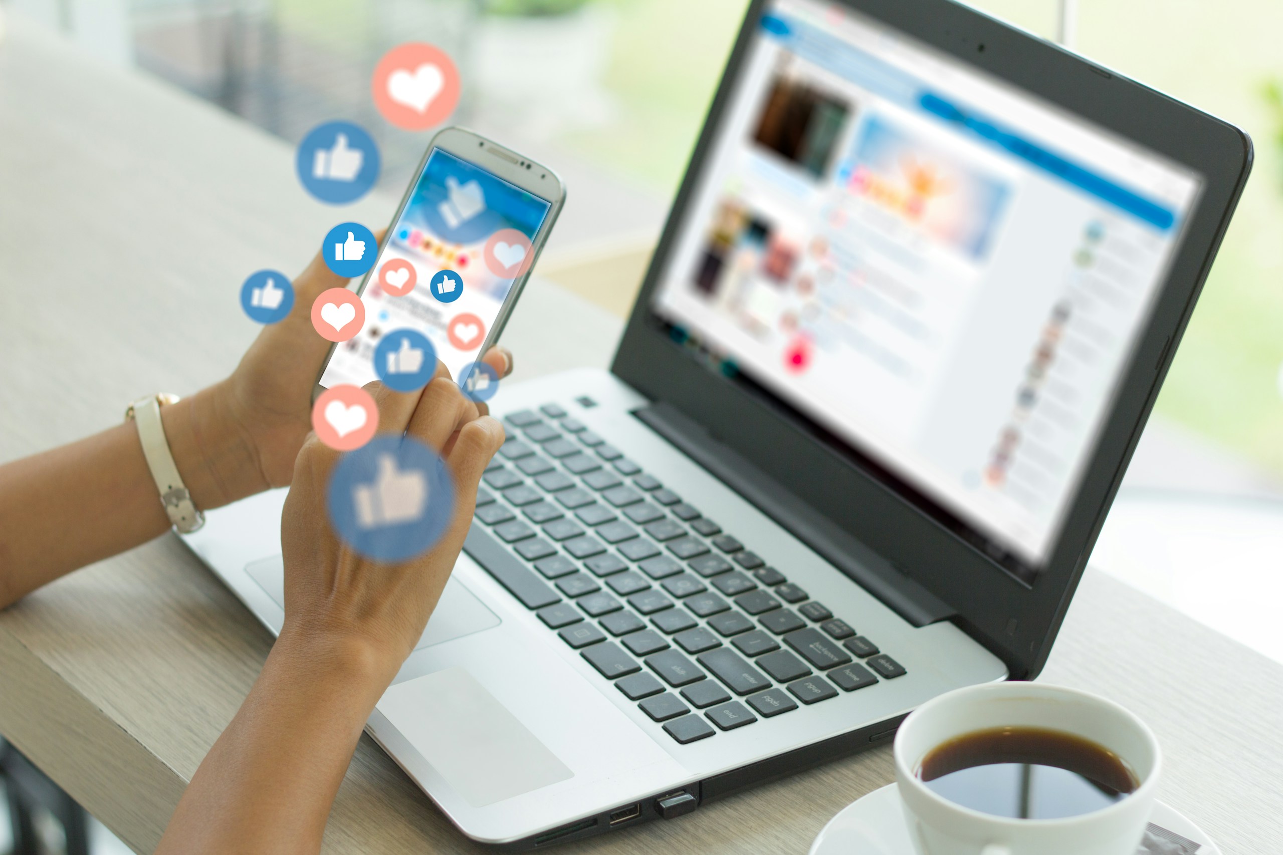 Frau mit Smartphone sitzt vor Laptop, fliegende Social Media Icons Likes und Herzen