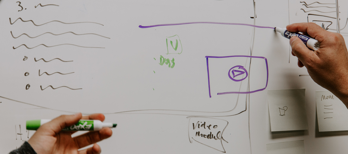 two people writing on a whiteboard with purple and green markers