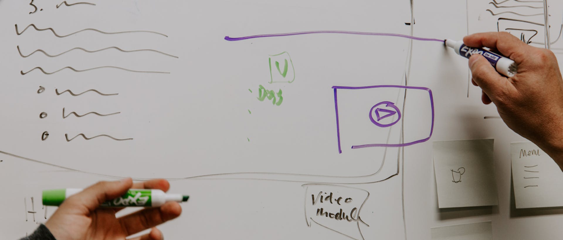 two people writing on a whiteboard with purple and green markers