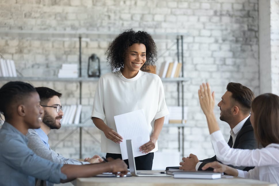 Junge afroamerikanische Frau steht lächelnd vor einem Tisch mit anderen Young Professionals