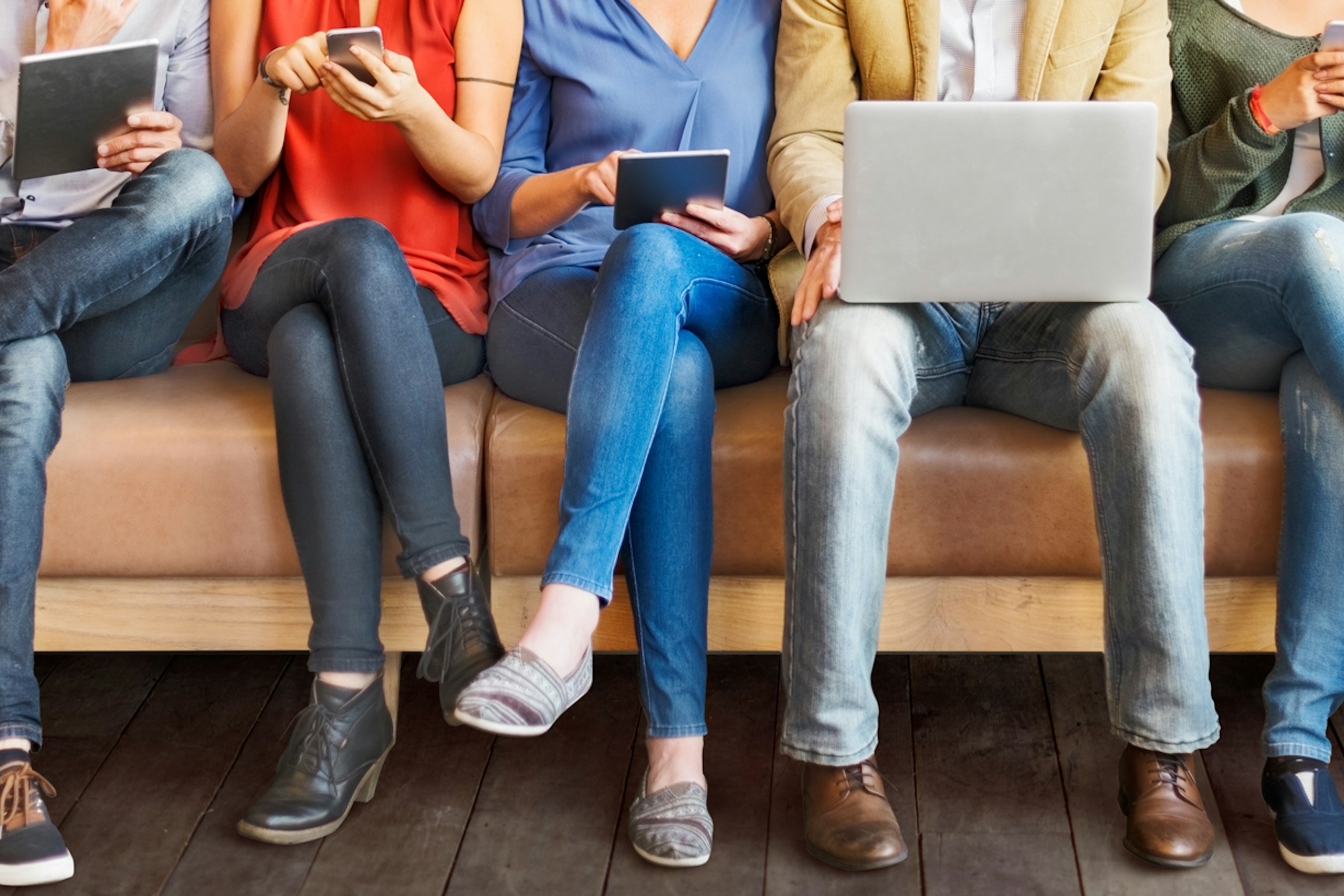 An image showing just the legs of several people sitting on a couch, on phones and laptops. Image for a guide to social media marketing