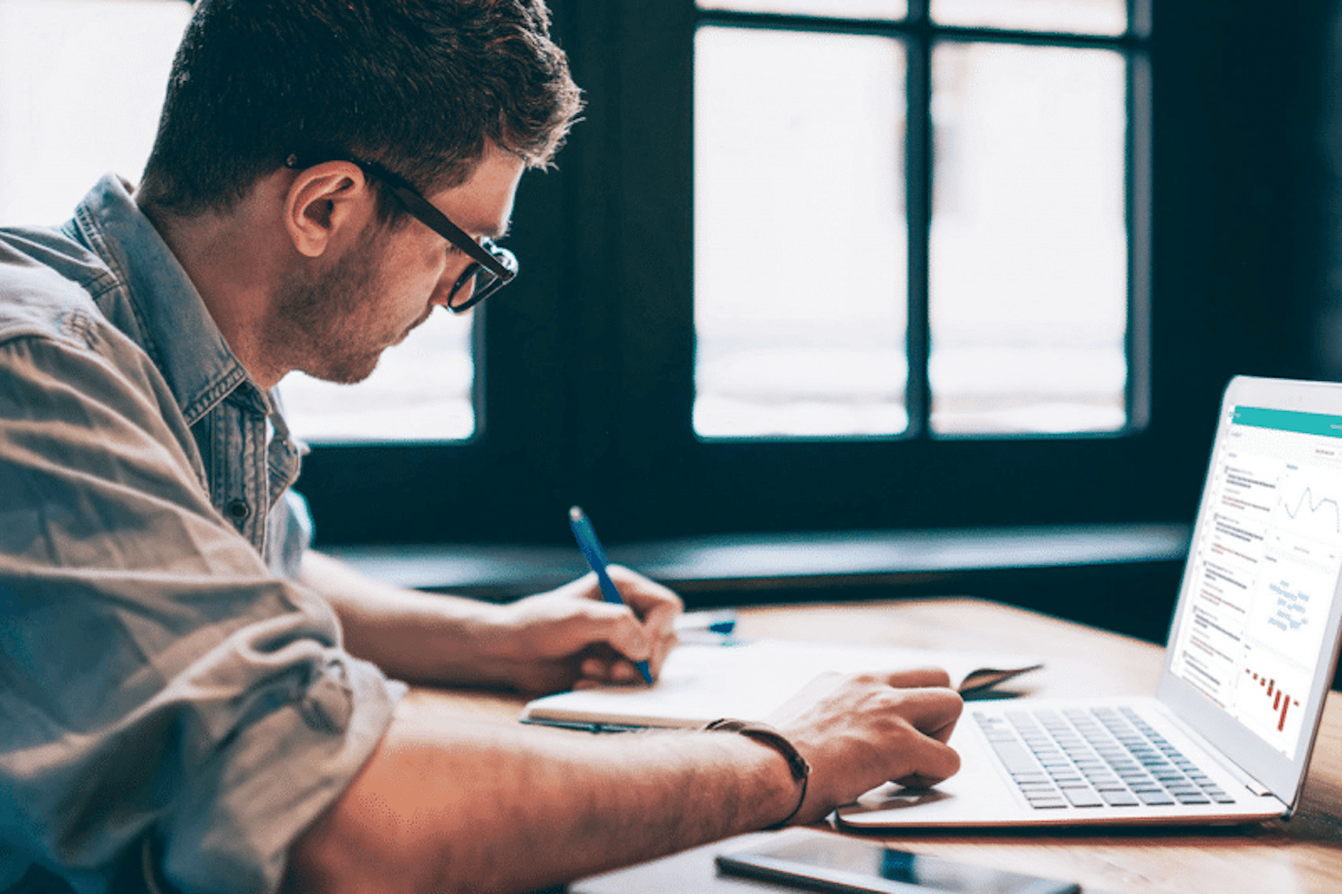 Man writing on a notepad while using the Meltwater platform on his laptop