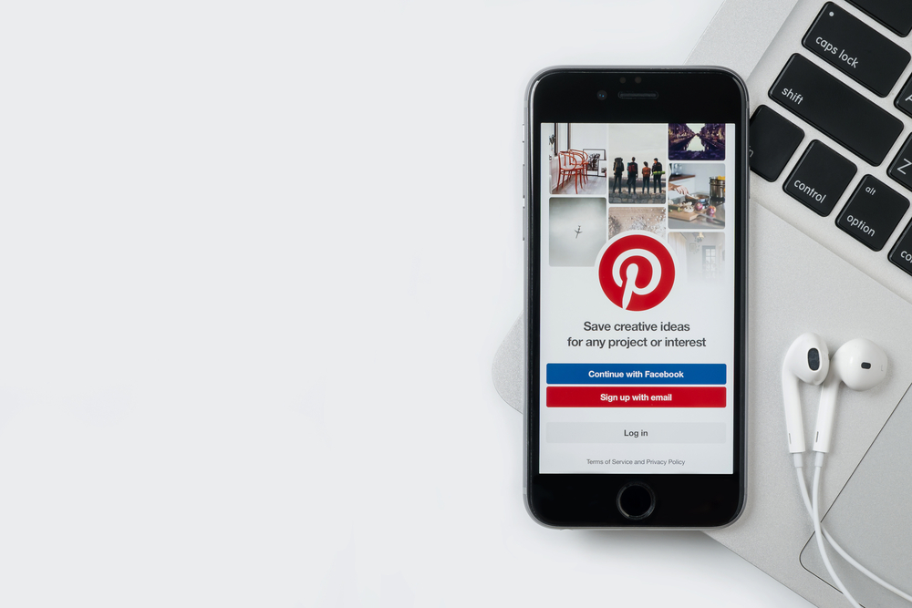 Un smartphone affichant la page de connexion Pinterest est posé sur un ordinateur portable à côté duquel on apperçoit des écouteurs