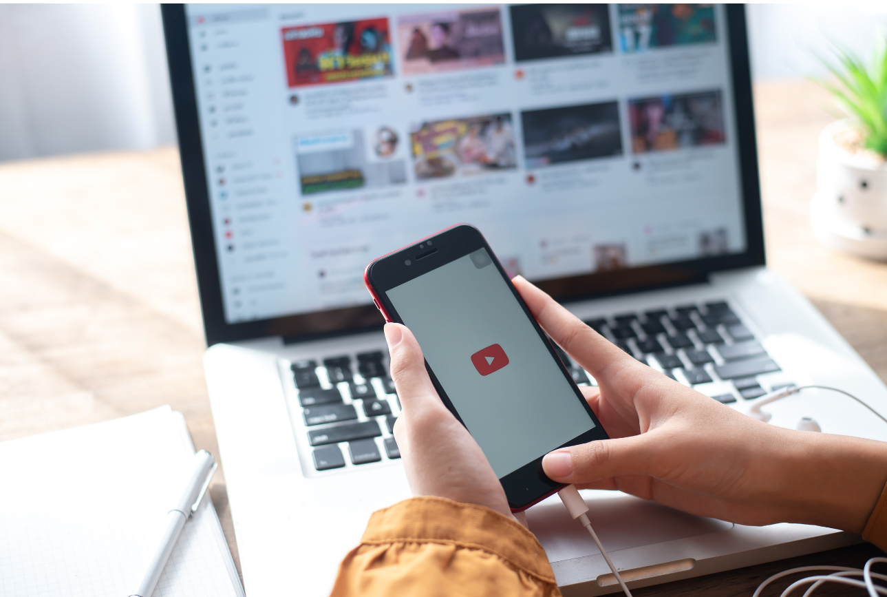 Une main tient un smartphone arborant un logo youtube sur fond blanc, derrière laquelle se trouve un ordinateur portable affichant la page d'accueil de youtube. A sa droite se trouve une plante verte et à sa gauche, un stylo posé sur un carnet. 