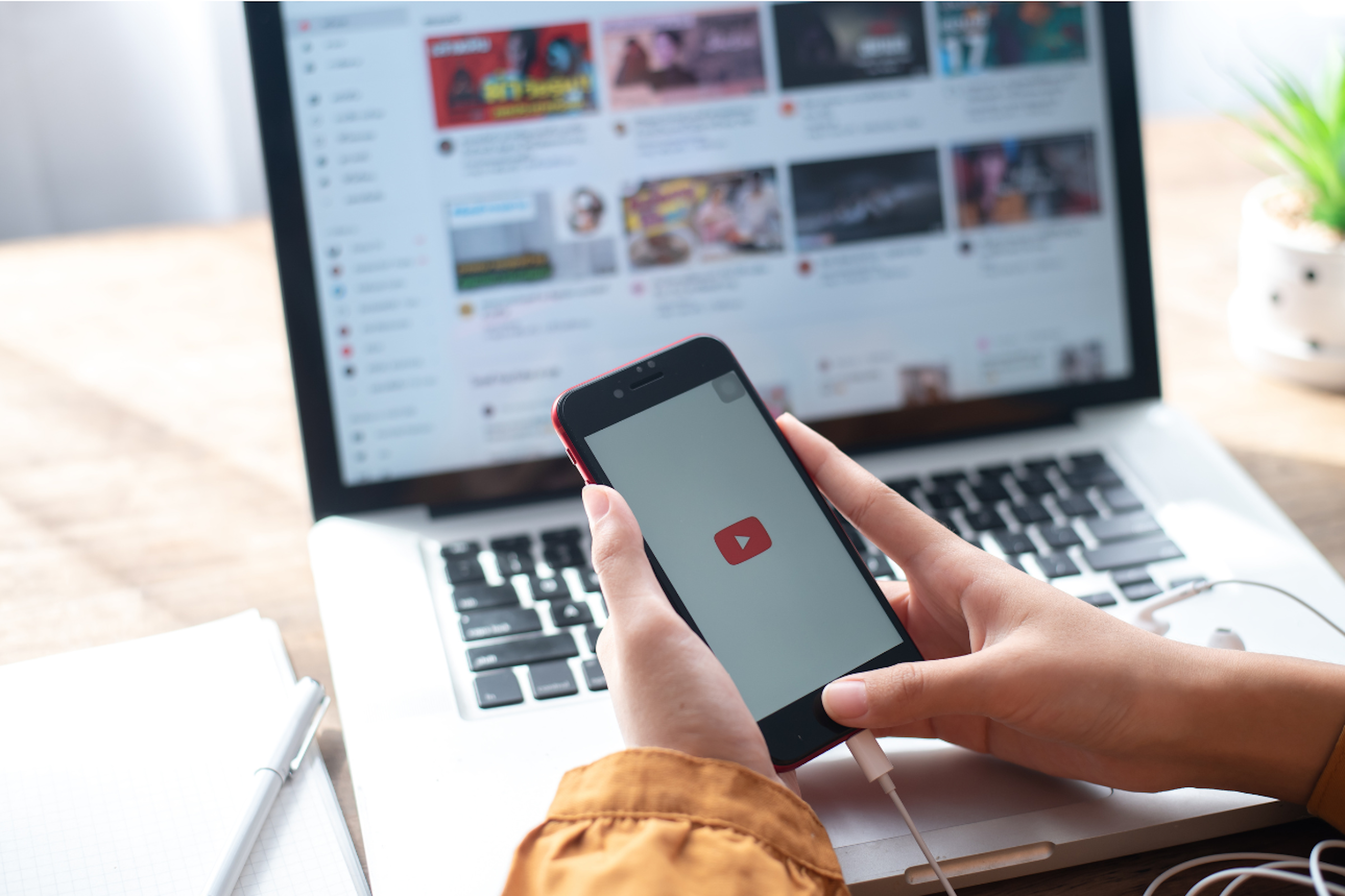 Une main tient un smartphone arborant un logo youtube sur fond blanc, derrière laquelle se trouve un ordinateur portable affichant la page d'accueil de youtube. A sa droite se trouve une plante verte et à sa gauche, un stylo posé sur un carnet.