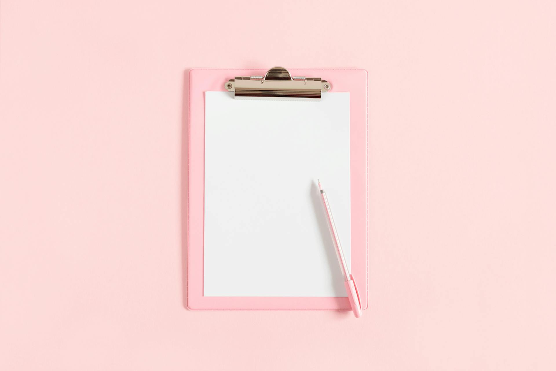 Clipboard with blank piece of paper and pen on light pink background. 10 examples of effective Press Releases.