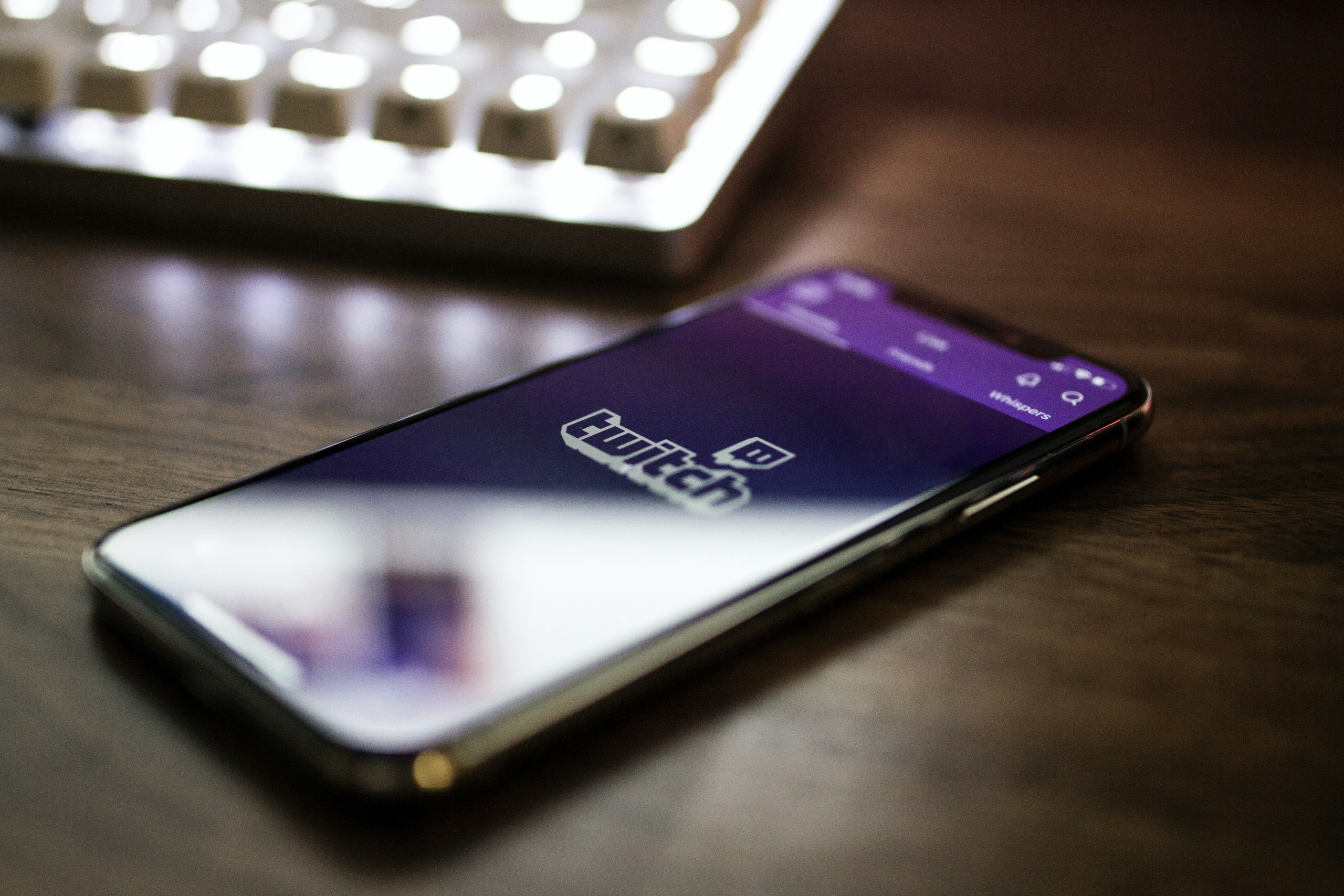 Un smartphone affichant la page d'arrivée de twitch est posé sur un table en bois sur laquelle on apperçoit également un clavier d'ordinateur.