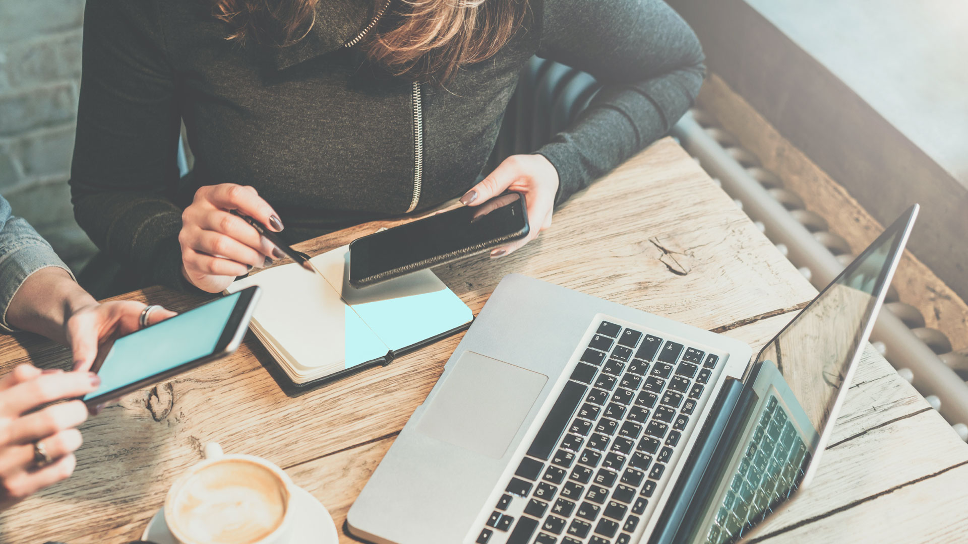A woman making notes in a notebook while looking at her smartphone and laptop screen