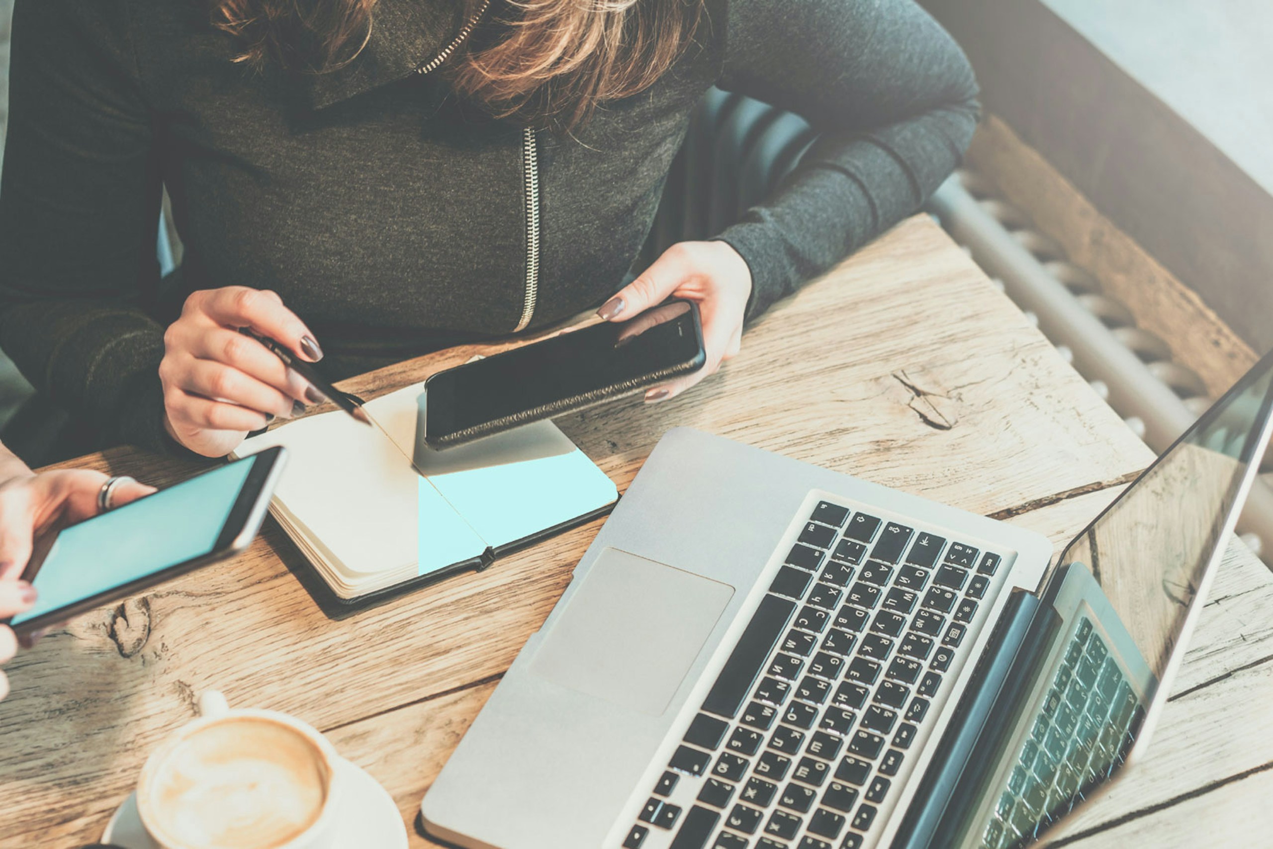 A woman making notes in a notebook while looking at her smartphone and laptop screen