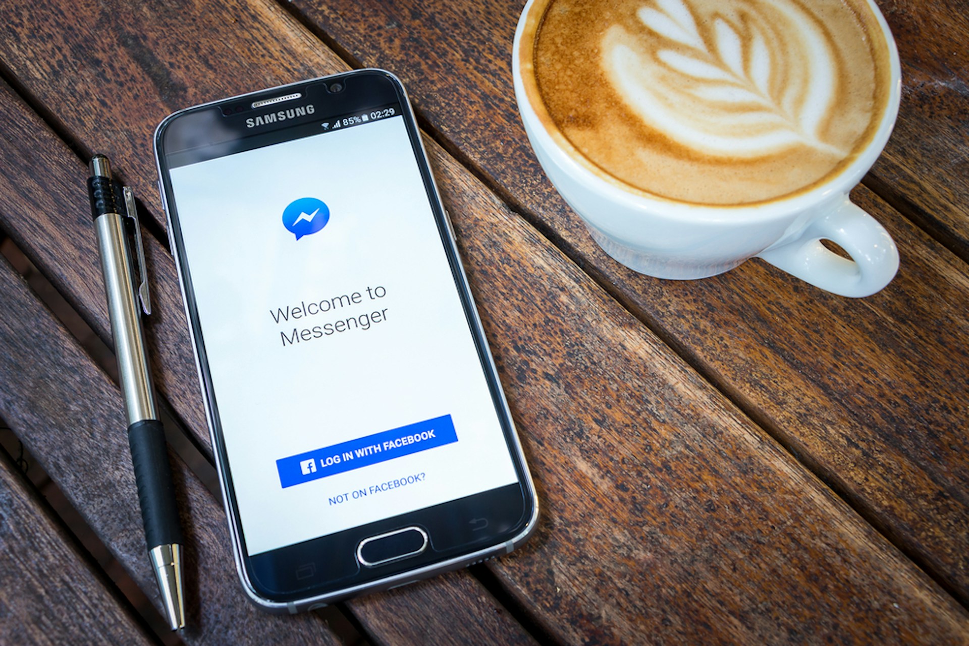 Man sieht ein Smartphone mit dem Facebook Messenger geöffnet auf einem Holztisch neben einer Tasse Kaffee liegen
