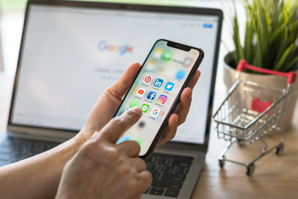 A hand scrolling through social icons on a smartphone while a Google search page is open on a laptop screen