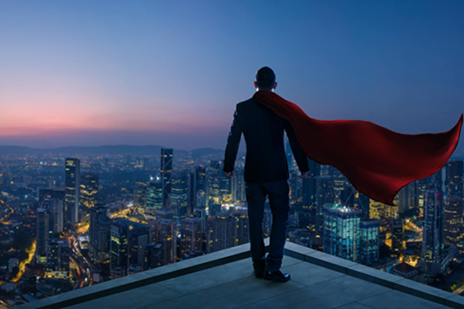 A man standing on top of a building looking over the city lights wearing a cape. 