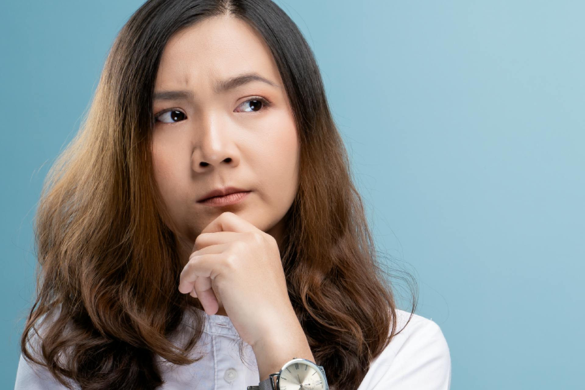 Young asian woman with her hand under her chin, listening, thinking, with a blue background.