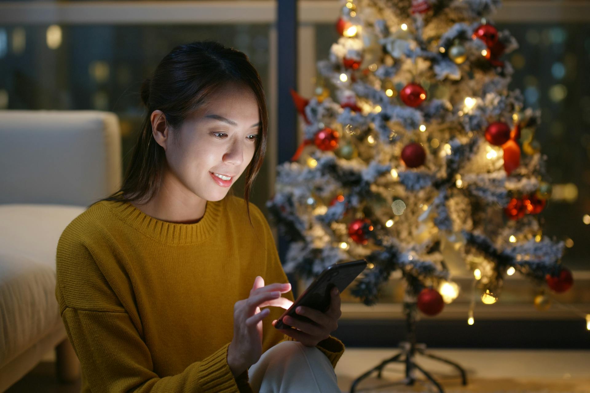 Junge asiatische Frau sitzt mit ihrem Smarthphone vor einem Weihnachtsbaum