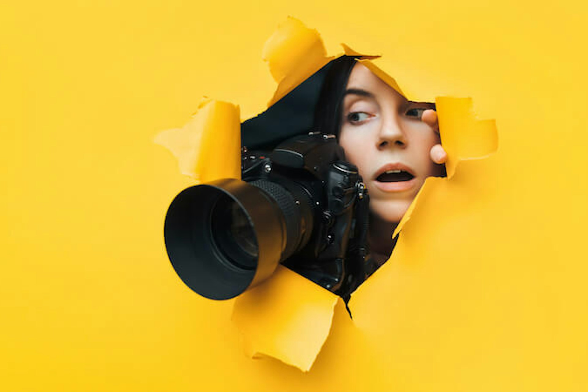 Man sieht eine junge Frau mit einer Kamera mit einem großen Objektiv durch eine gelbe Pappwand luken. Das Bild ist Header für unseren Beitrag zu elektronischen Pressespiegeln.
