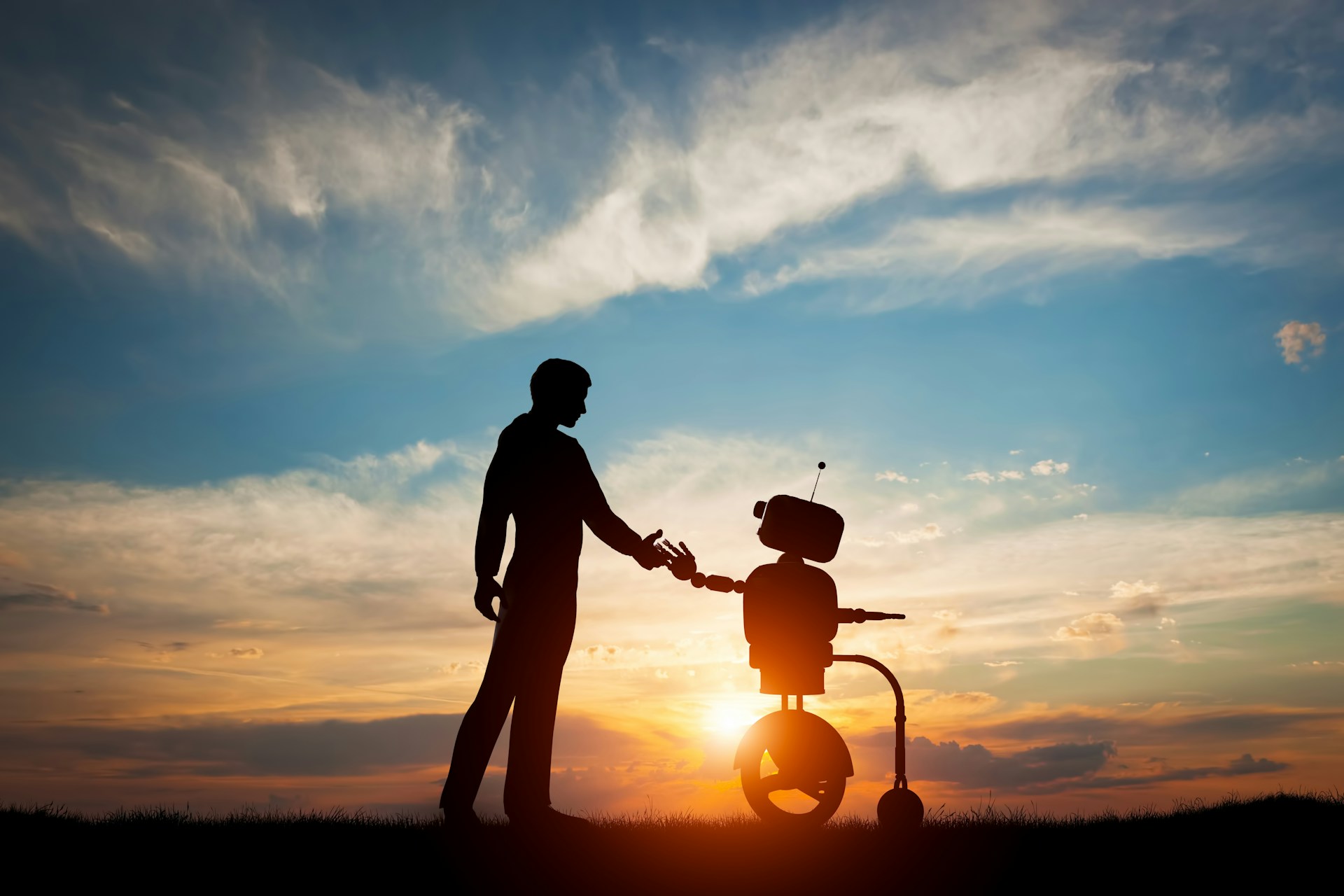 A man holding a robot's hand whilst standing in front of a sunset.