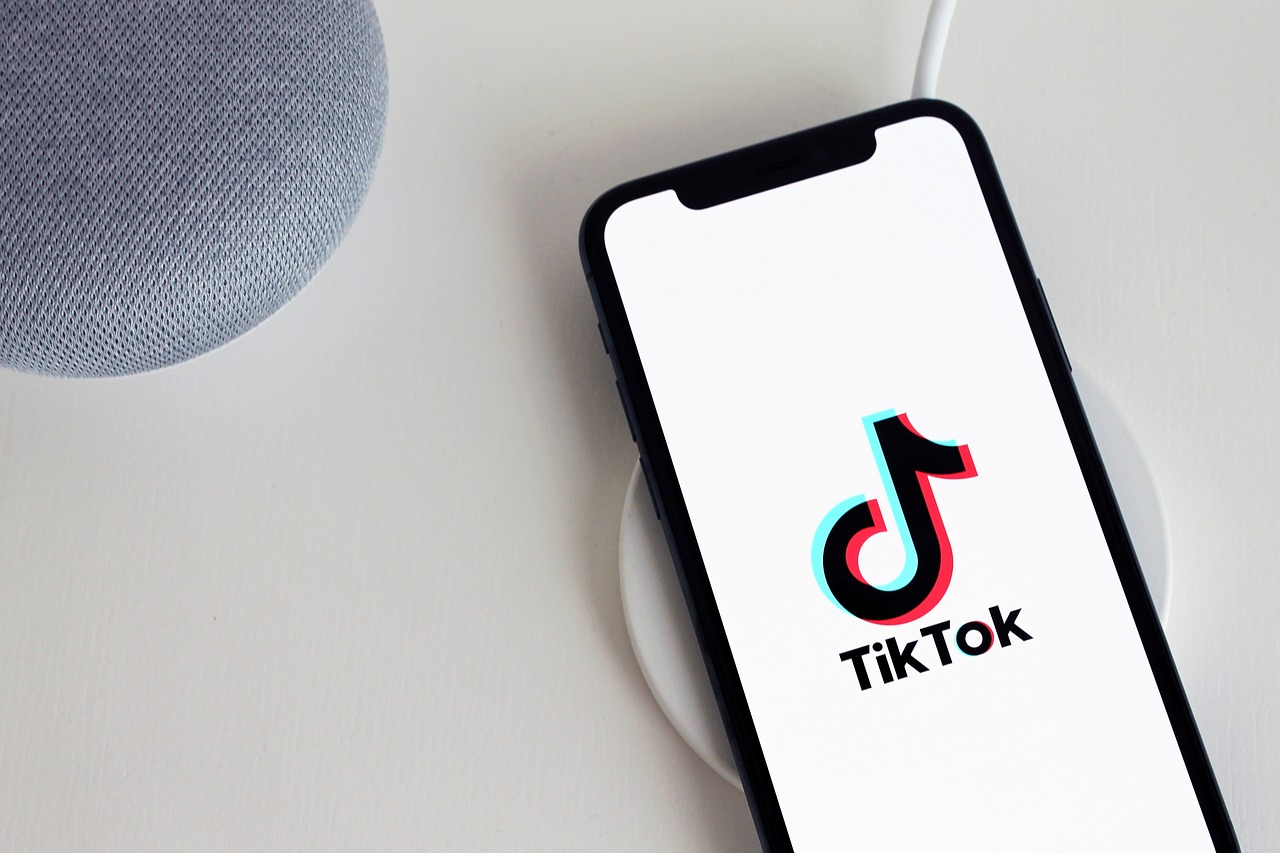 Un smartphone est disposé sur une base de recharge blanche à côté d'une enceinte grise. L'écran affiche le logo tiktok