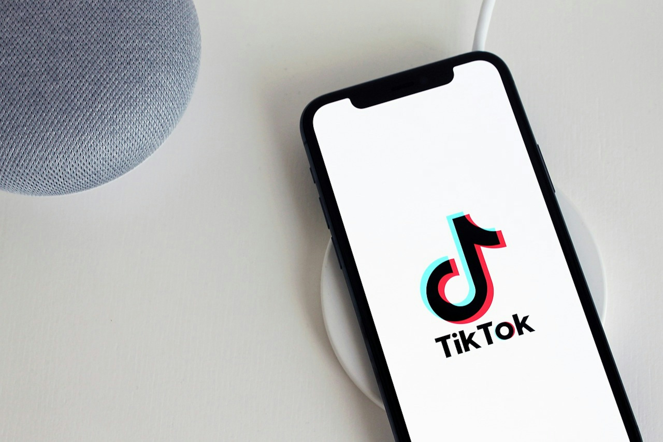 Un smartphone est disposé sur une base de recharge blanche à côté d'une enceinte grise. L'écran affiche le logo tiktok