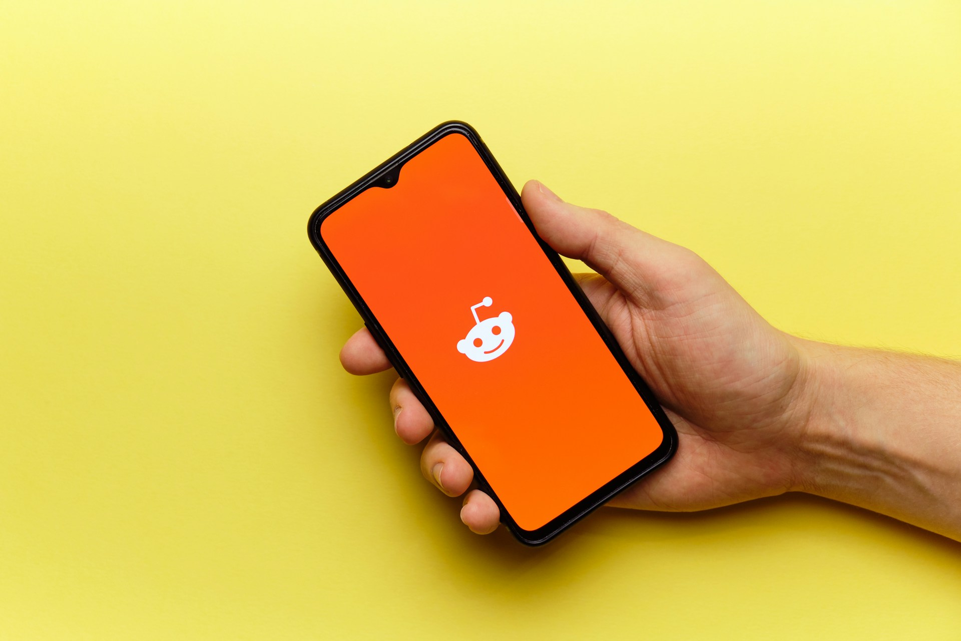 Auf einem gelben Hintergrund hält eine Hand ein Handy auf dem Mann das weiße Reddit-Symbol auf orangenen Hintergrund sieht