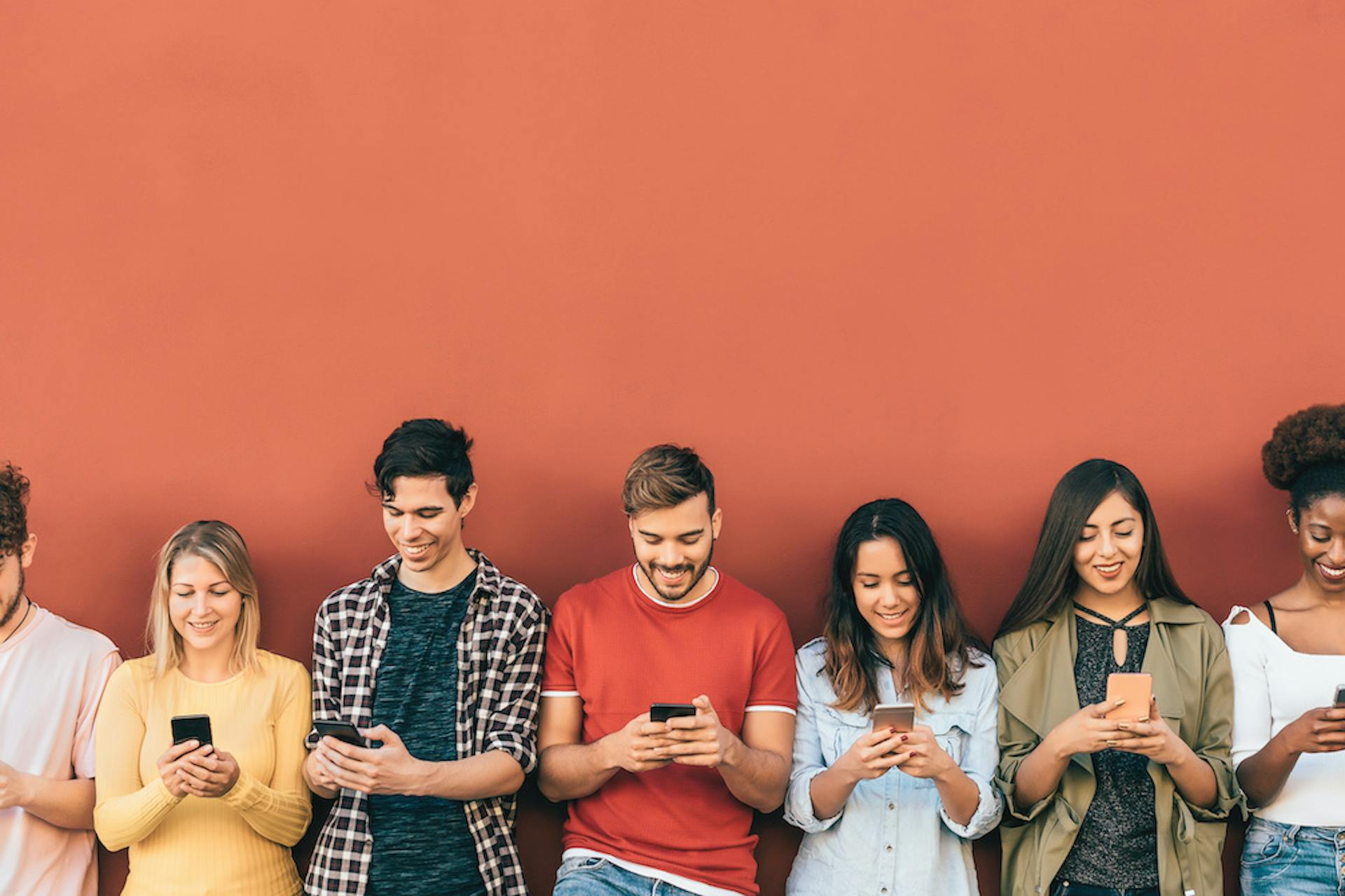 Junge Leute schauen fröhlich auf ihr Handy während sie an einer orangen Wand lehnen