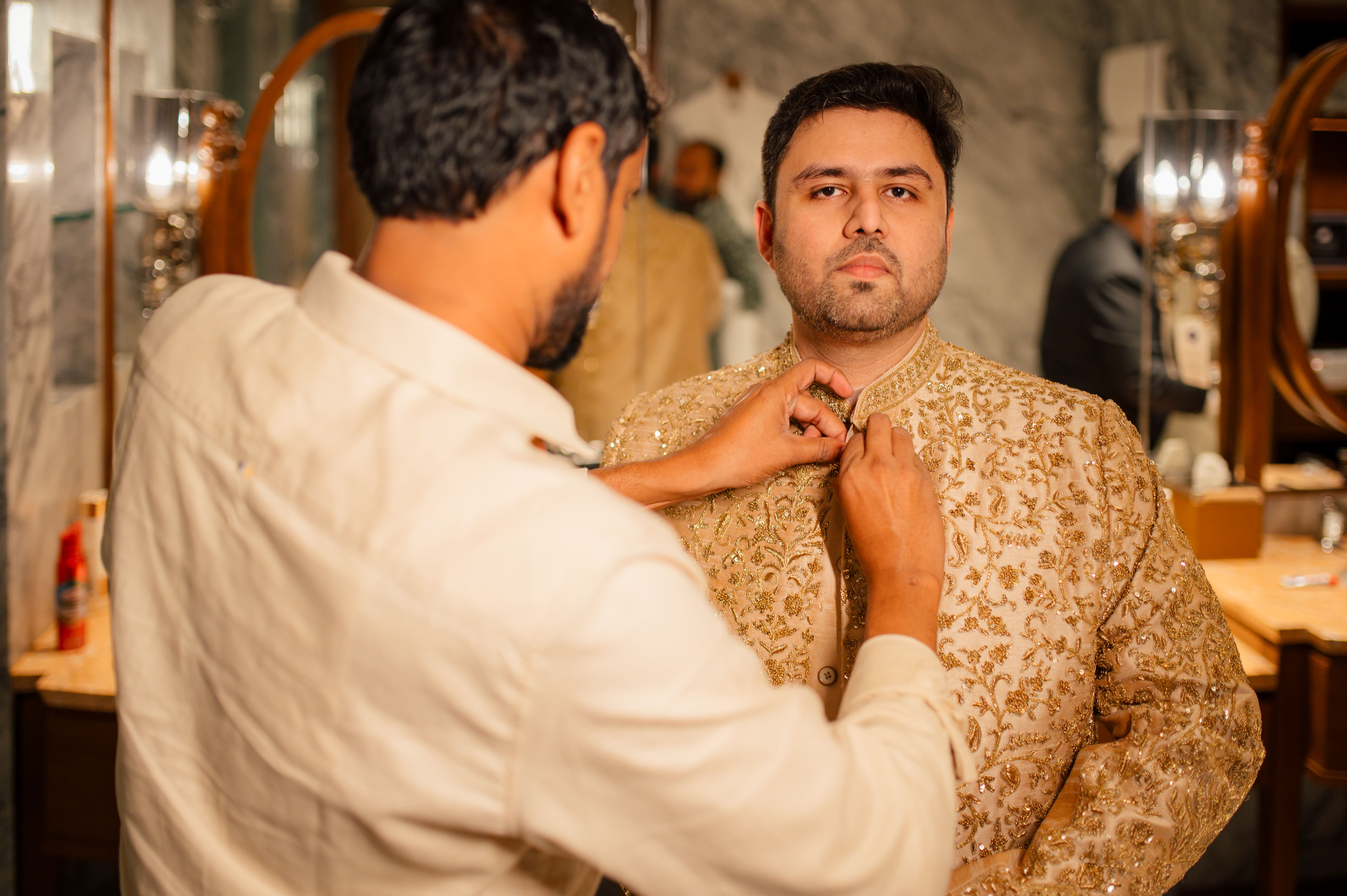 Groom getting dressed for wedding while his best friend is helping him tie his sherwani button