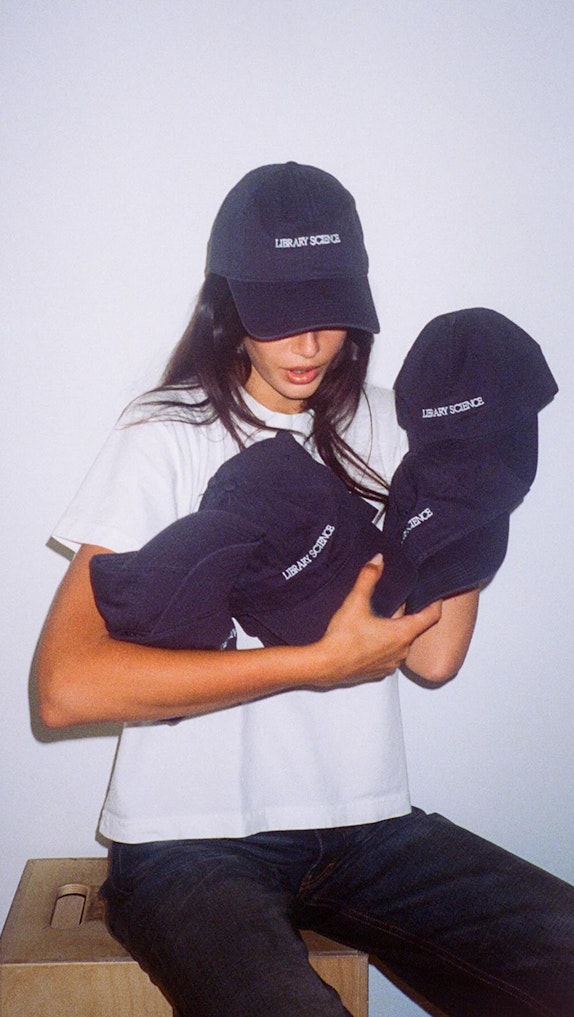 Model wearing navy "Library Science" cap while holding armful of matching caps, custom merchandise by MerchCamp