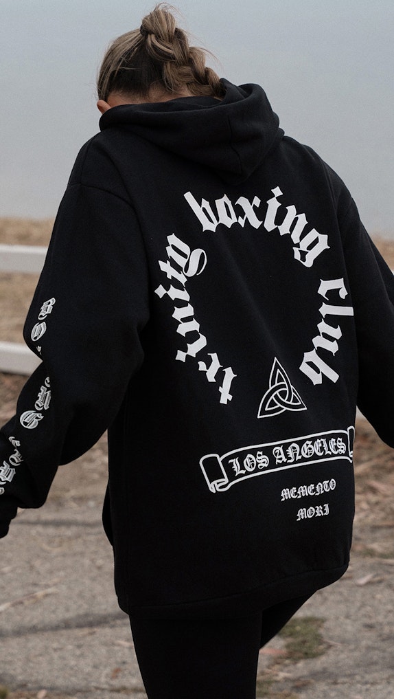 Woman wearing black oversized hoodie with Trinity Boxing Club Los Angeles gothic text design outdoors, custom merchandise by MerchCamp