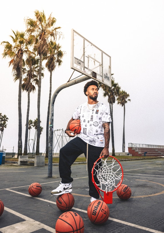 Man holding basketball and hoop rim on outdoor LA court surrounded by Wilson basketballs and palm trees — MerchCamp custom merchandise