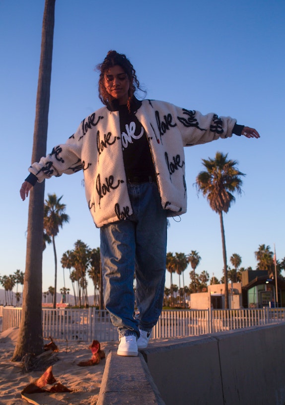 Woman in "love" print sherpa jacket balancing on beach wall with palm trees at golden hour — MerchCamp custom merchandise