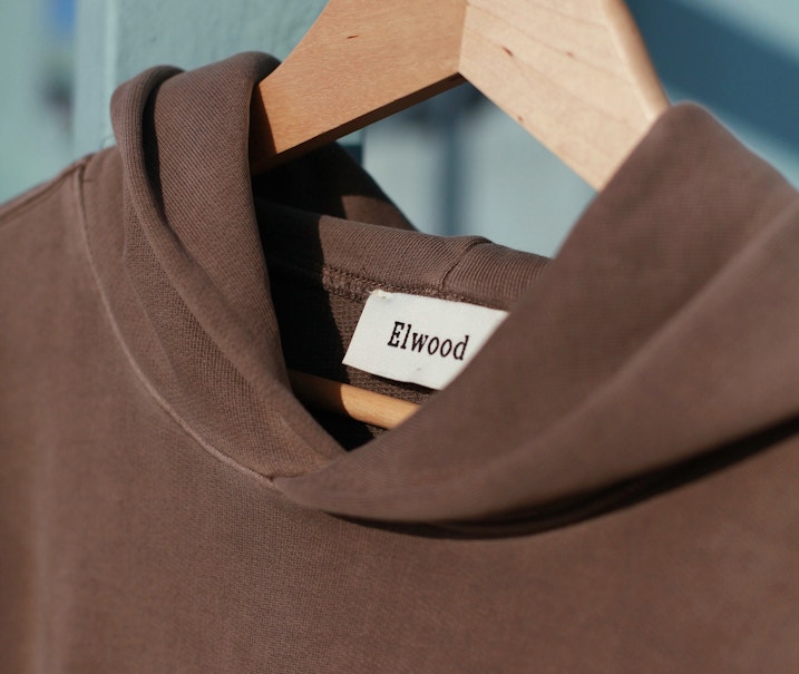 Brown Elwood brand t-shirt on wooden hanger showing neck label close-up — MerchCamp custom merchandise