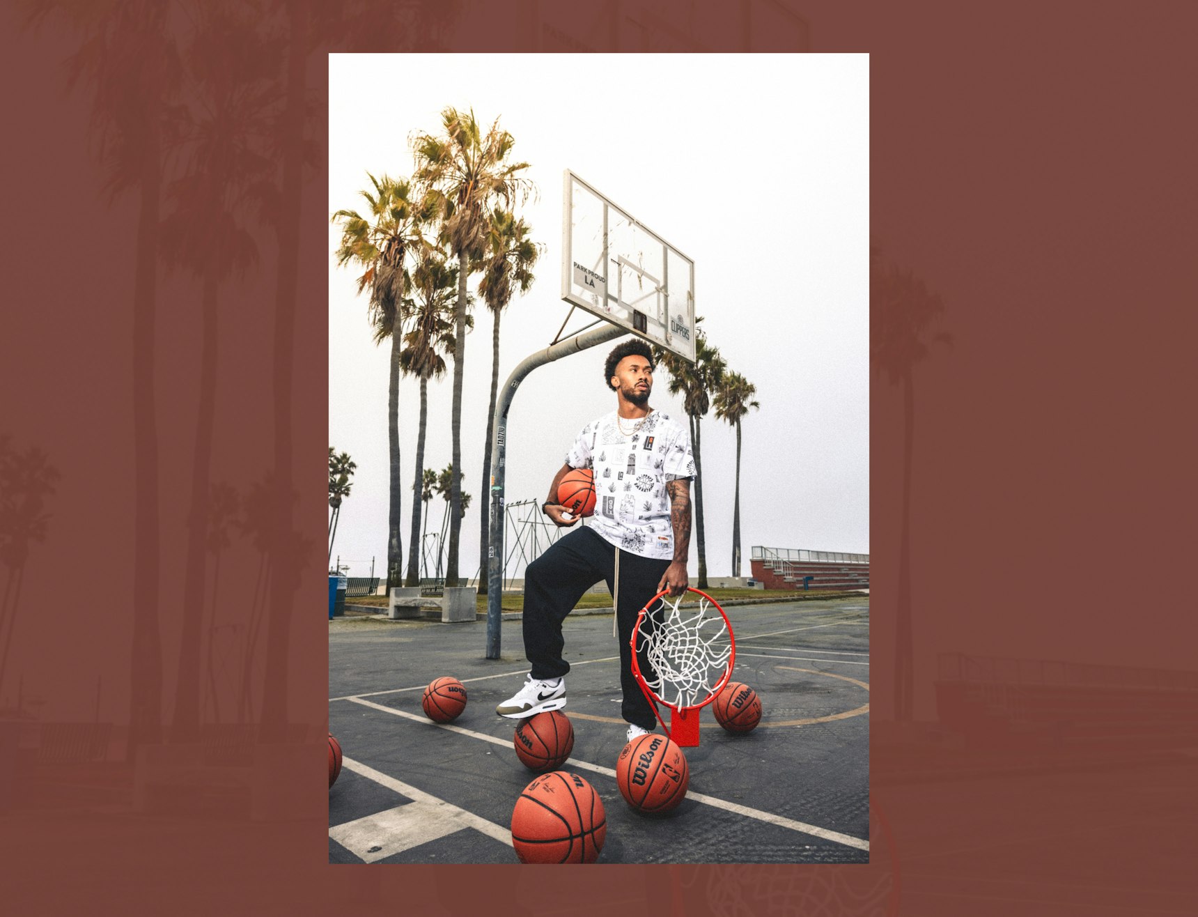Man posing on outdoor basketball court with Wilson basketballs, basketball rim, and palm trees — LA Clippers merchandise by MerchCamp